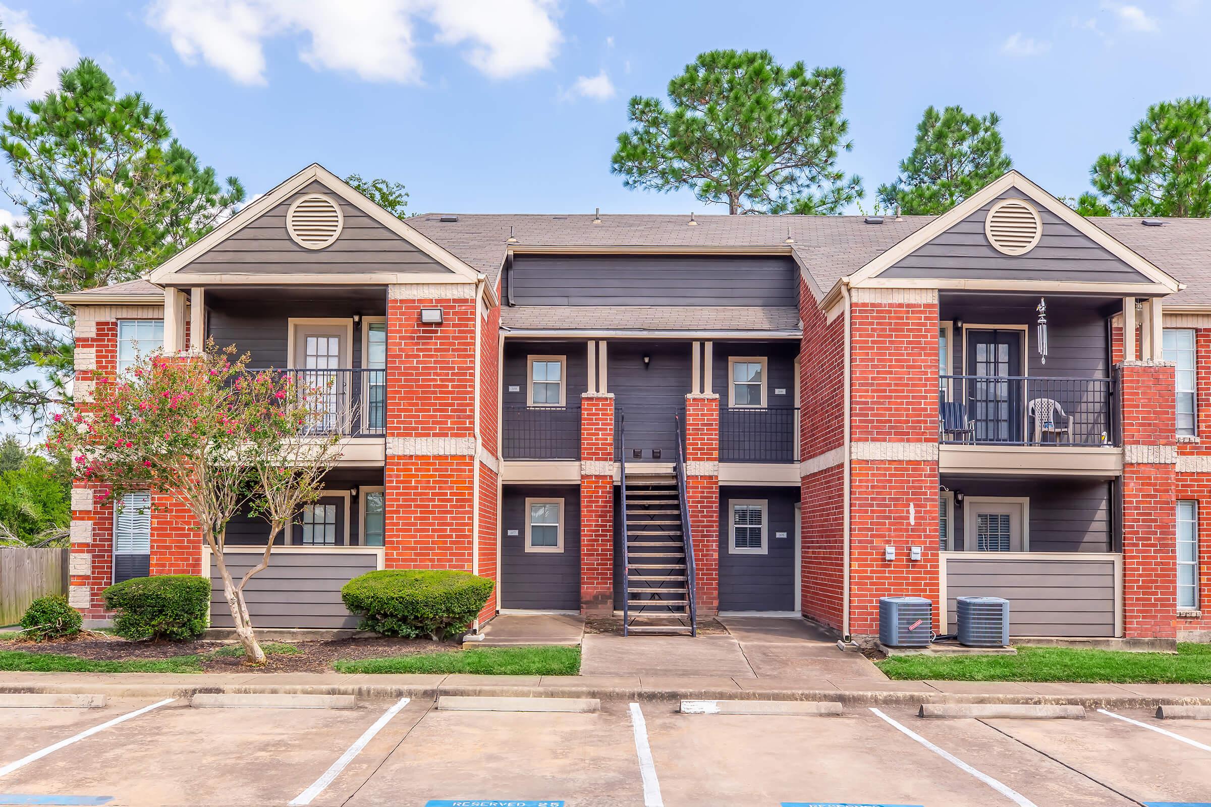 Exterior view of a two-story apartment building featuring red brick and dark siding. Balconies are visible with outdoor furniture. Lush green trees frame the scene, and parking spaces are located in the foreground. The sky is clear with some clouds.