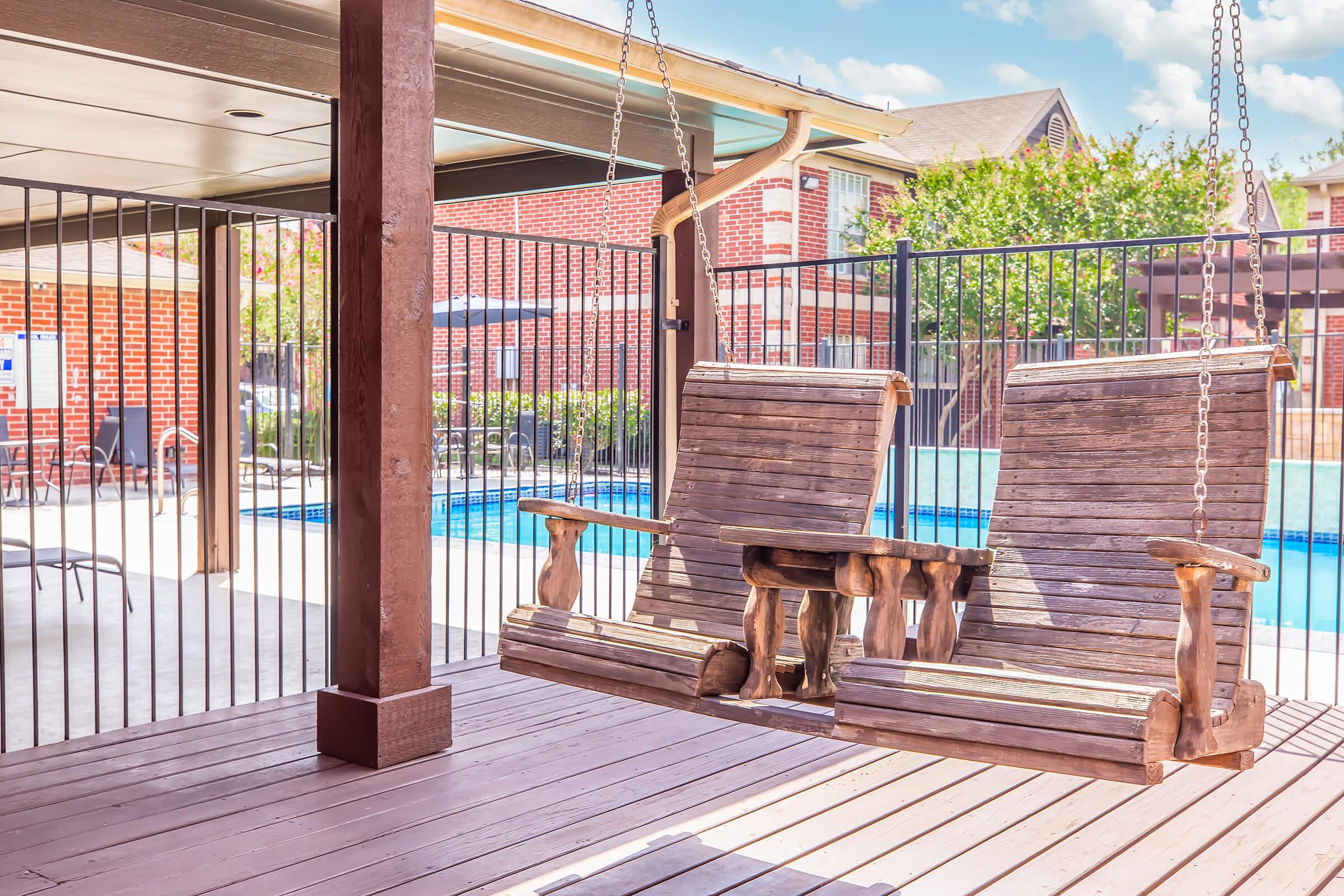 A wooden swing set with two seats, suspended from a covered patio, overlooking a swimming pool area. The surrounding fence is visible, and the scene features sunny weather with clear blue skies and green trees in the background.