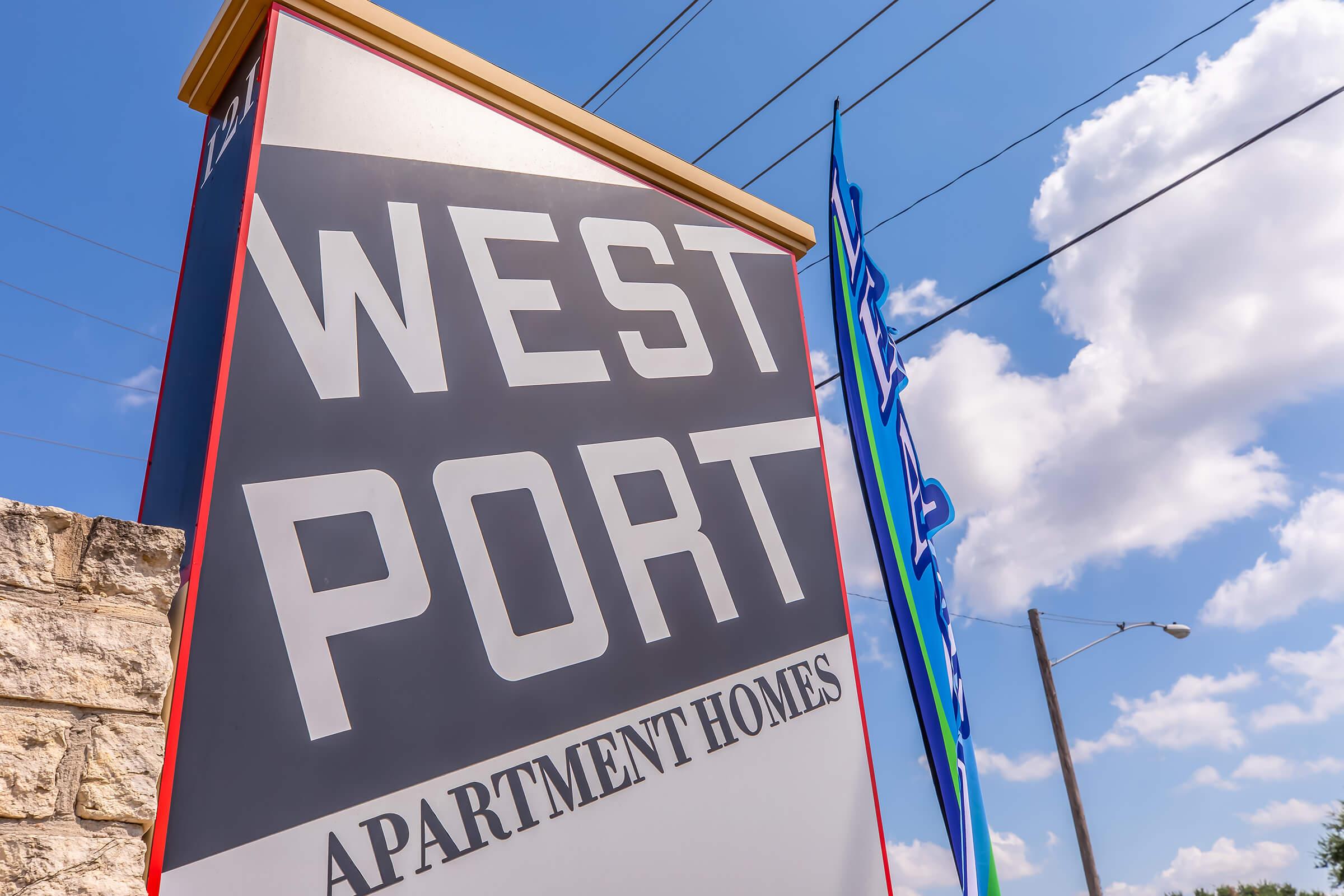 Sign for West Port Apartment Homes, featuring large bold lettering against a clear blue sky with fluffy clouds. The sign also includes colorful flags nearby, indicating the location of the apartments.
