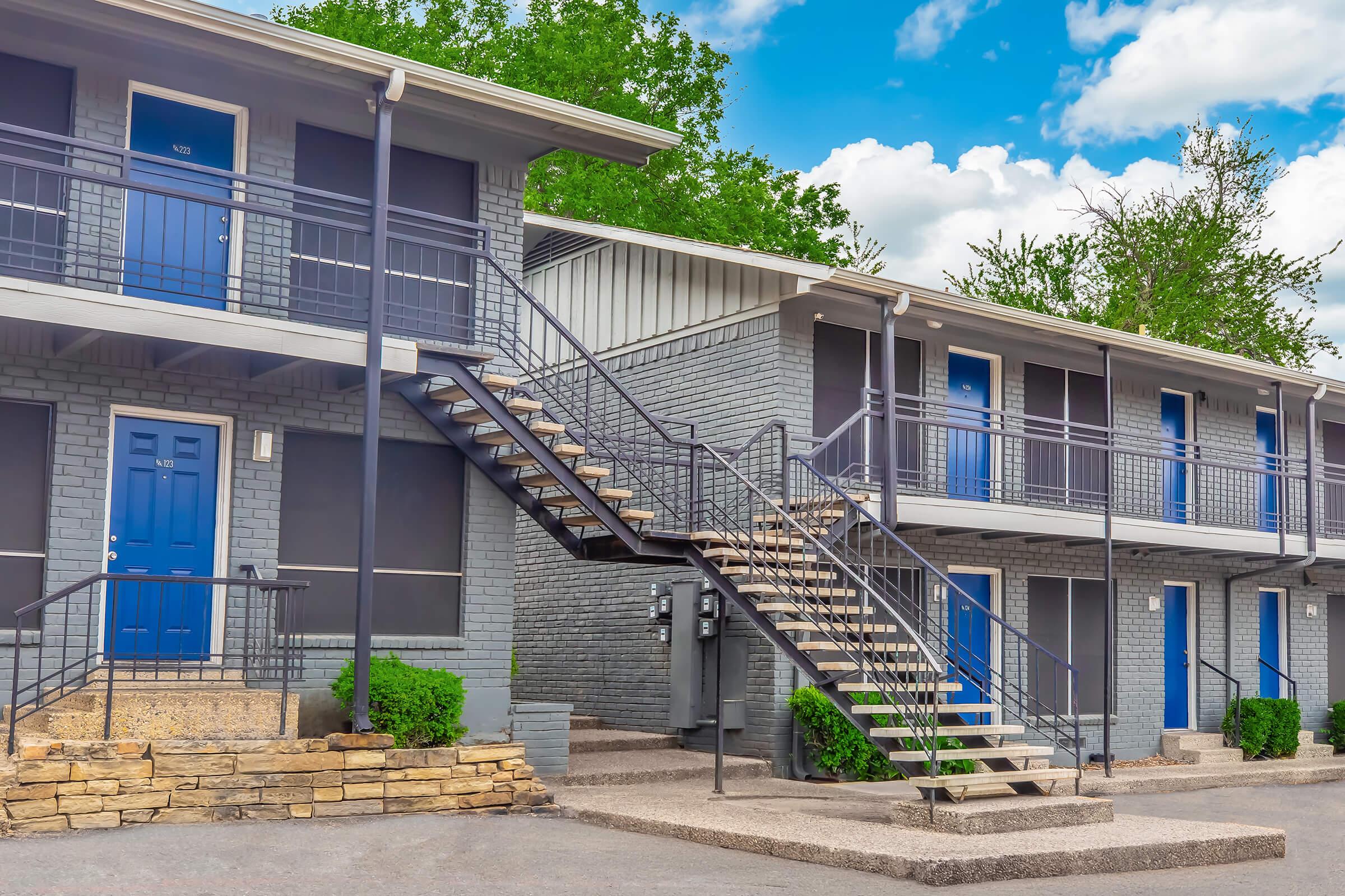 A two-story building with gray exterior walls and blue doors, featuring a staircase leading to the upper level. The entrance has a landscaped area with bushes, and the sky above is partly cloudy, adding a bright and inviting atmosphere.