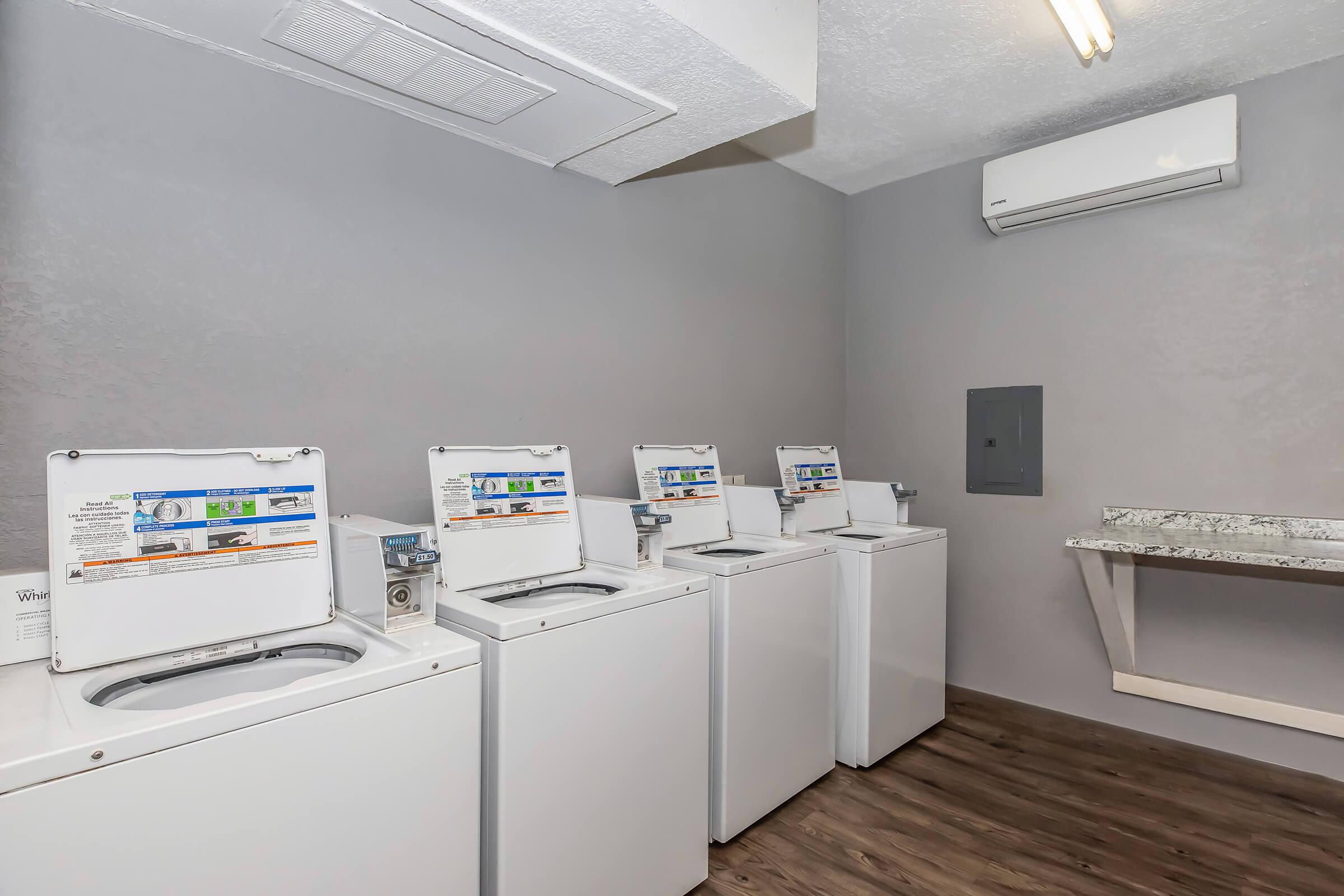 A laundry room featuring four white washing machines aligned in a row, with informational stickers on each. There is a countertop and an air conditioning unit on the wall, with a light fixture overhead. The floor is wooden, and the walls are painted gray, creating a clean and functional space.