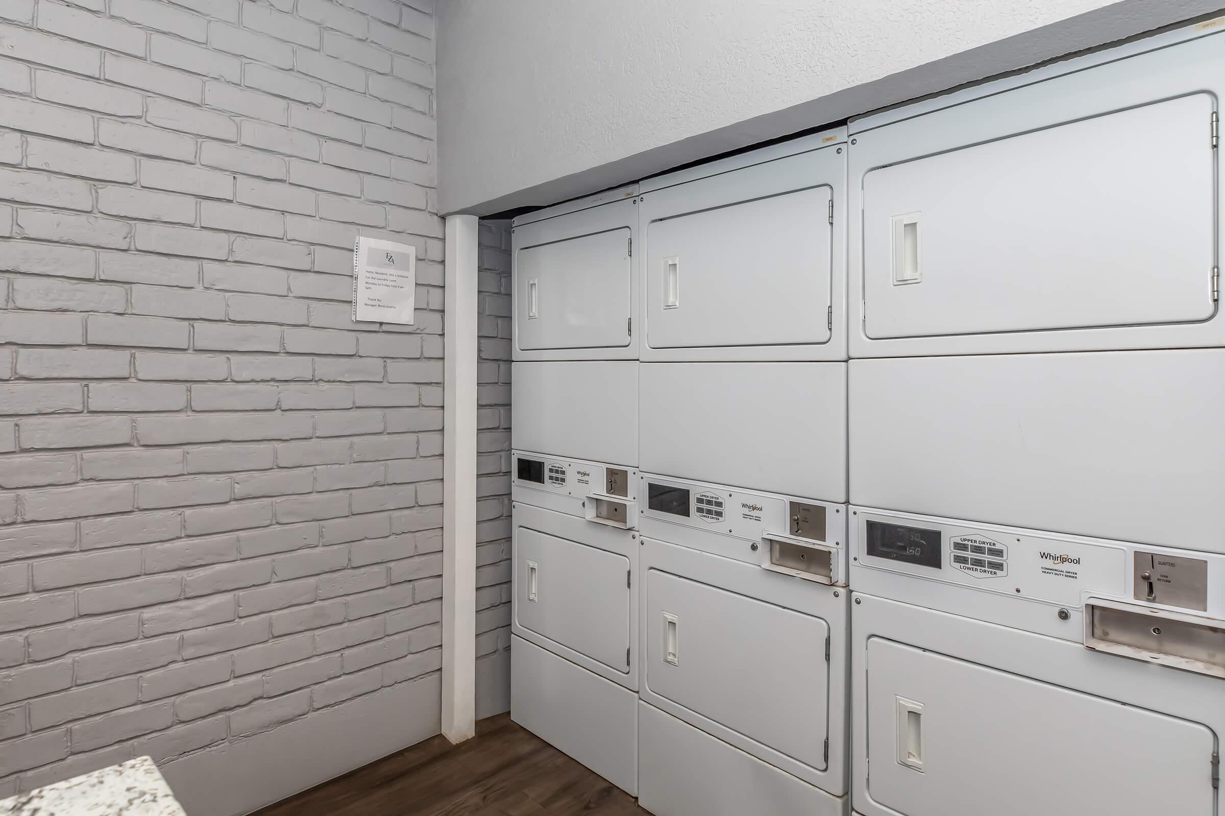 A row of white laundry lockers against a light gray brick wall, featuring electronic controls and slots for securing items. The area is well-lit, with a modern design and a small informational sign on the wall beside the lockers.
