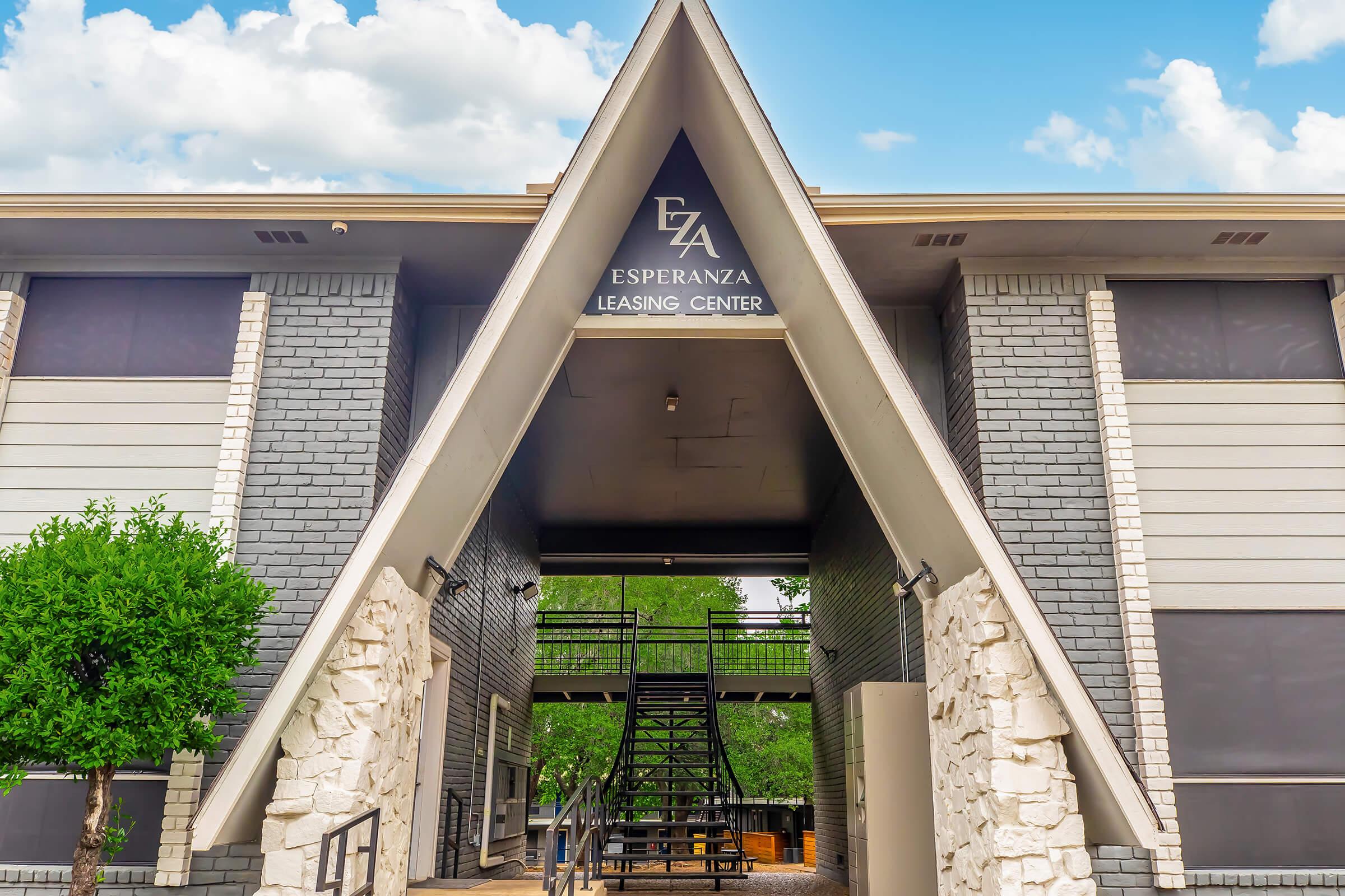Exterior view of the Esperanza Leasing Center featuring an A-frame architectural design. The entrance is framed by stone accents and surrounded by greenery, under a clear blue sky with scattered clouds. A staircase leads up to the leasing office, emphasizing a welcoming atmosphere.