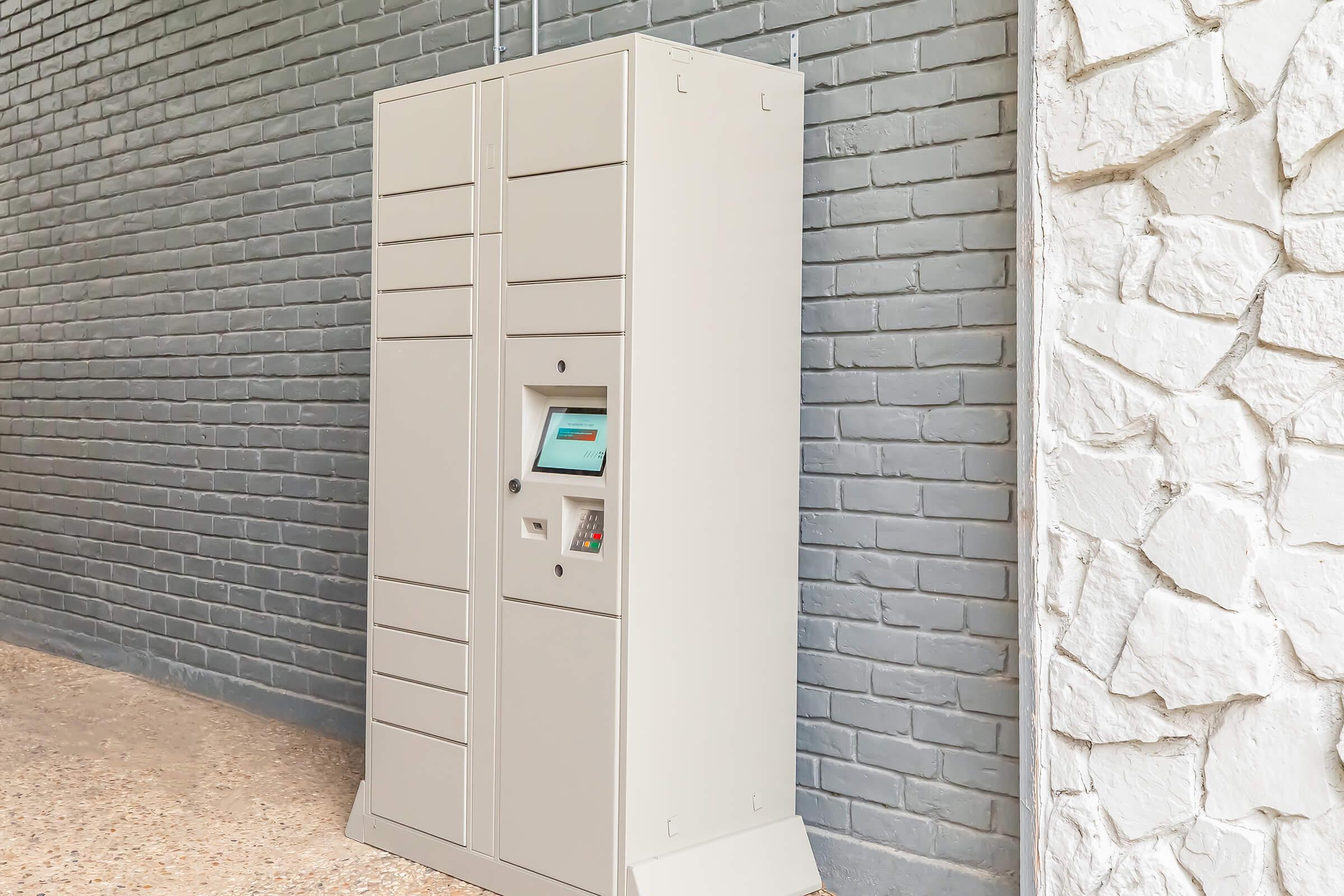 A modern, gray package delivery locker unit against a textured wall. The locker has multiple compartments for secure package storage and features a digital interface for user interaction. The area around the locker is clean and well-maintained.