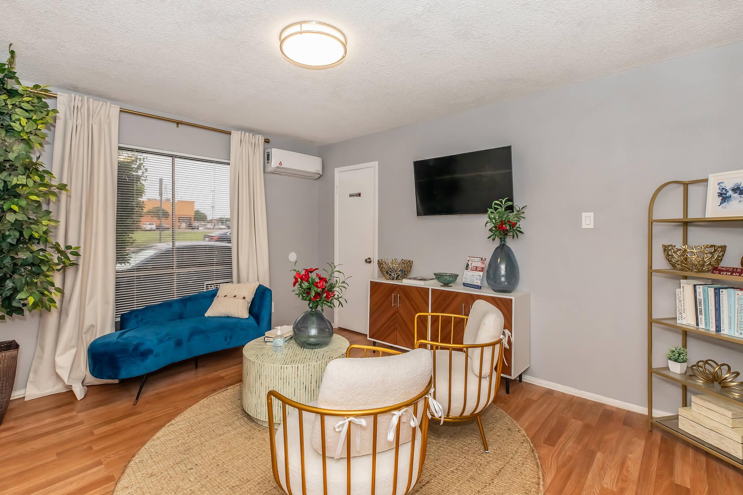 A cozy living room featuring a blue velvet sofa, two round chairs with golden frames, a small coffee table, and a decorative rug. There’s a flat-screen TV mounted on the wall, large curtains, and several houseplants. A sideboard holds books and decorative items, while a vase with red flowers adds a pop of color.