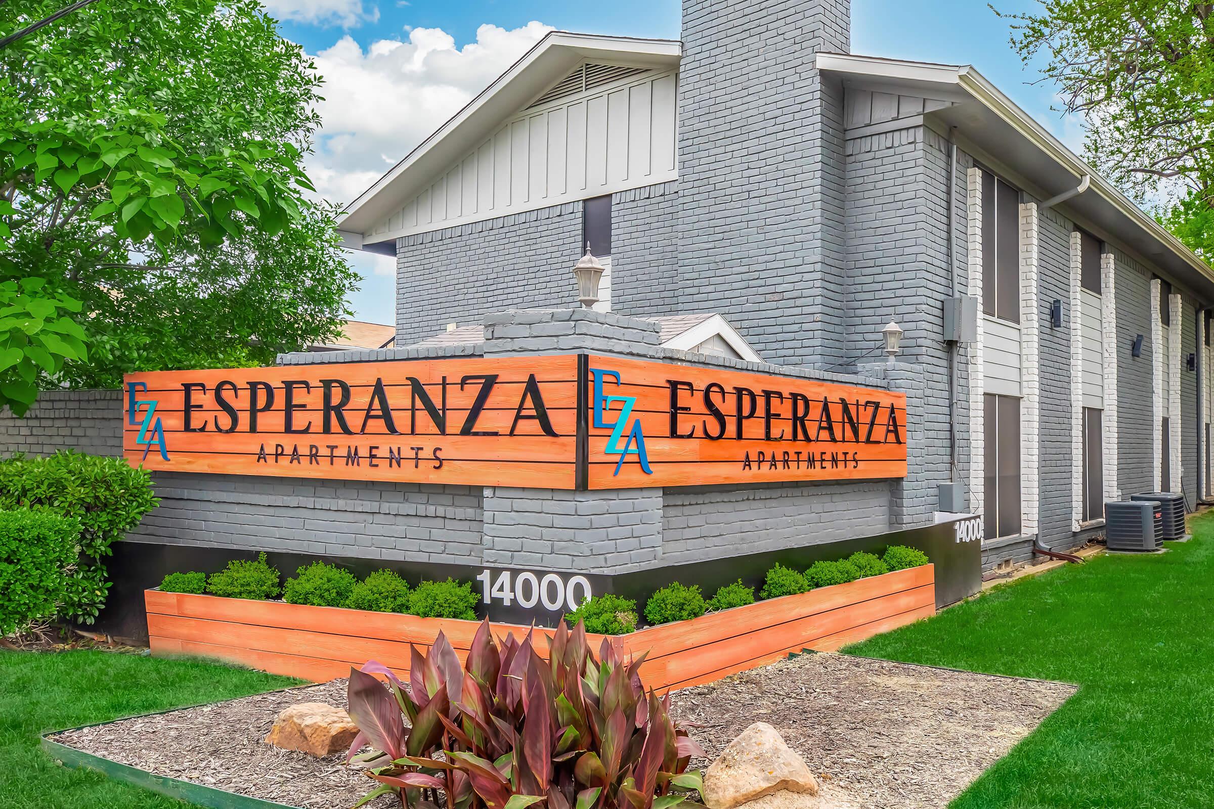 A well-maintained apartment building featuring a large sign that reads "Esperanza Apartments." The sign is prominently displayed with a modern design against a backdrop of green grass and landscaping, including plants and rocks, under a clear blue sky.