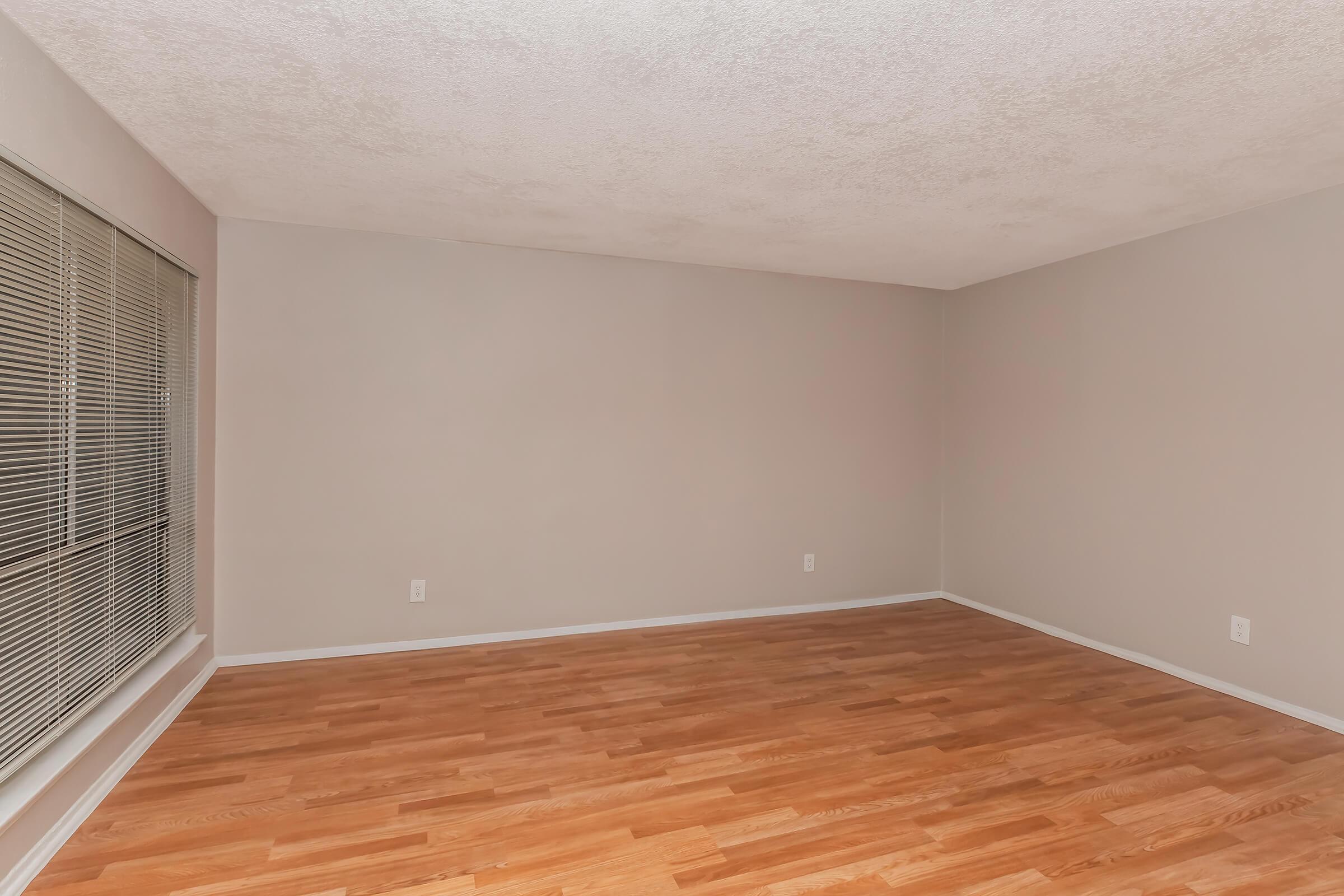 A spacious, empty room with light-colored walls and hardwood flooring. One wall features large windows with horizontal blinds, allowing natural light to enter. The room has a simple and uncluttered layout, creating a calm and neutral atmosphere.