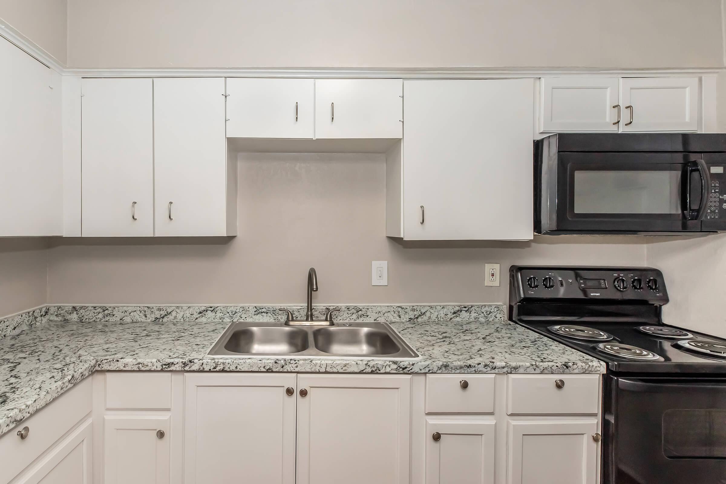 A modern kitchen featuring white cabinets, a granite countertop, a double sink with a sleek faucet, and stainless steel appliances including a microwave and a stovetop. The walls are painted a light color, providing a clean and bright atmosphere.