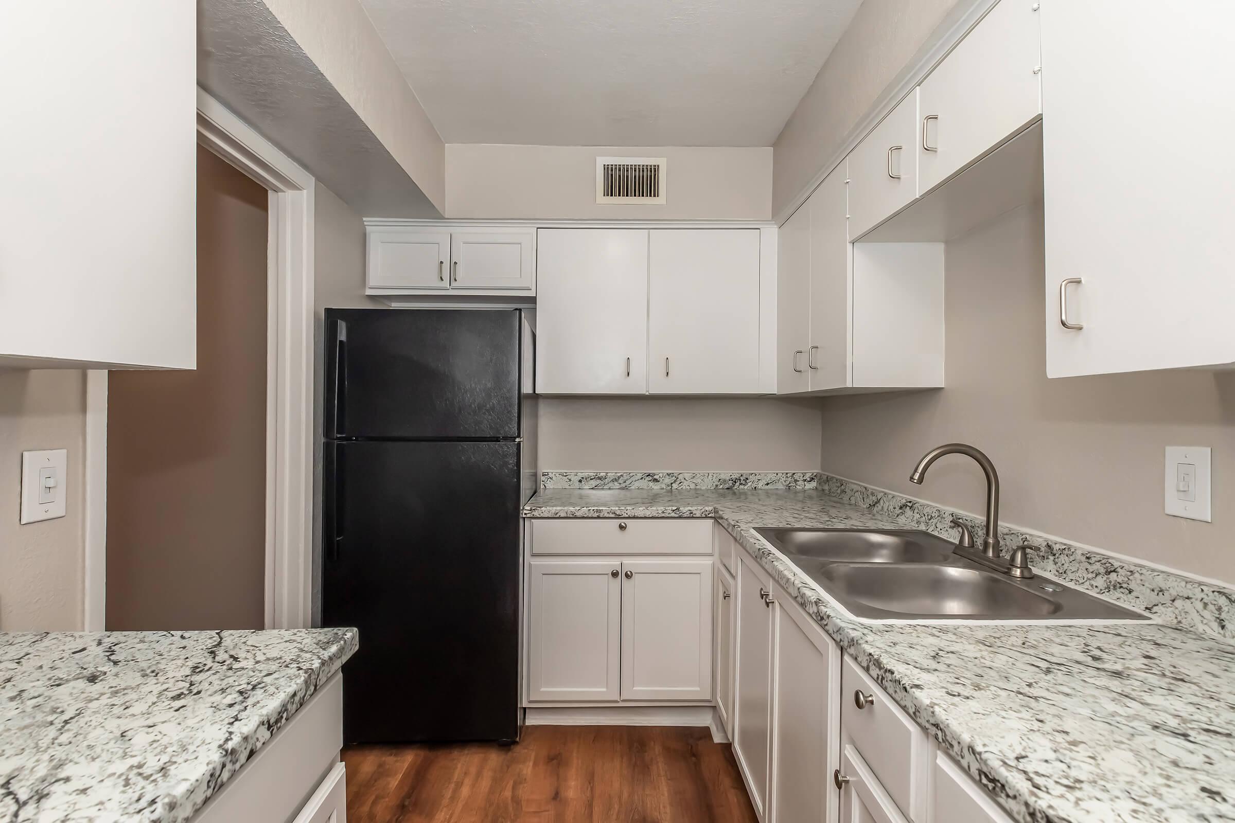 A modern kitchen featuring white cabinets, a black refrigerator, and dual sinks. The countertops are made of a speckled gray stone, and there is a wood floor. The walls are painted in a light color, creating a bright and inviting atmosphere.