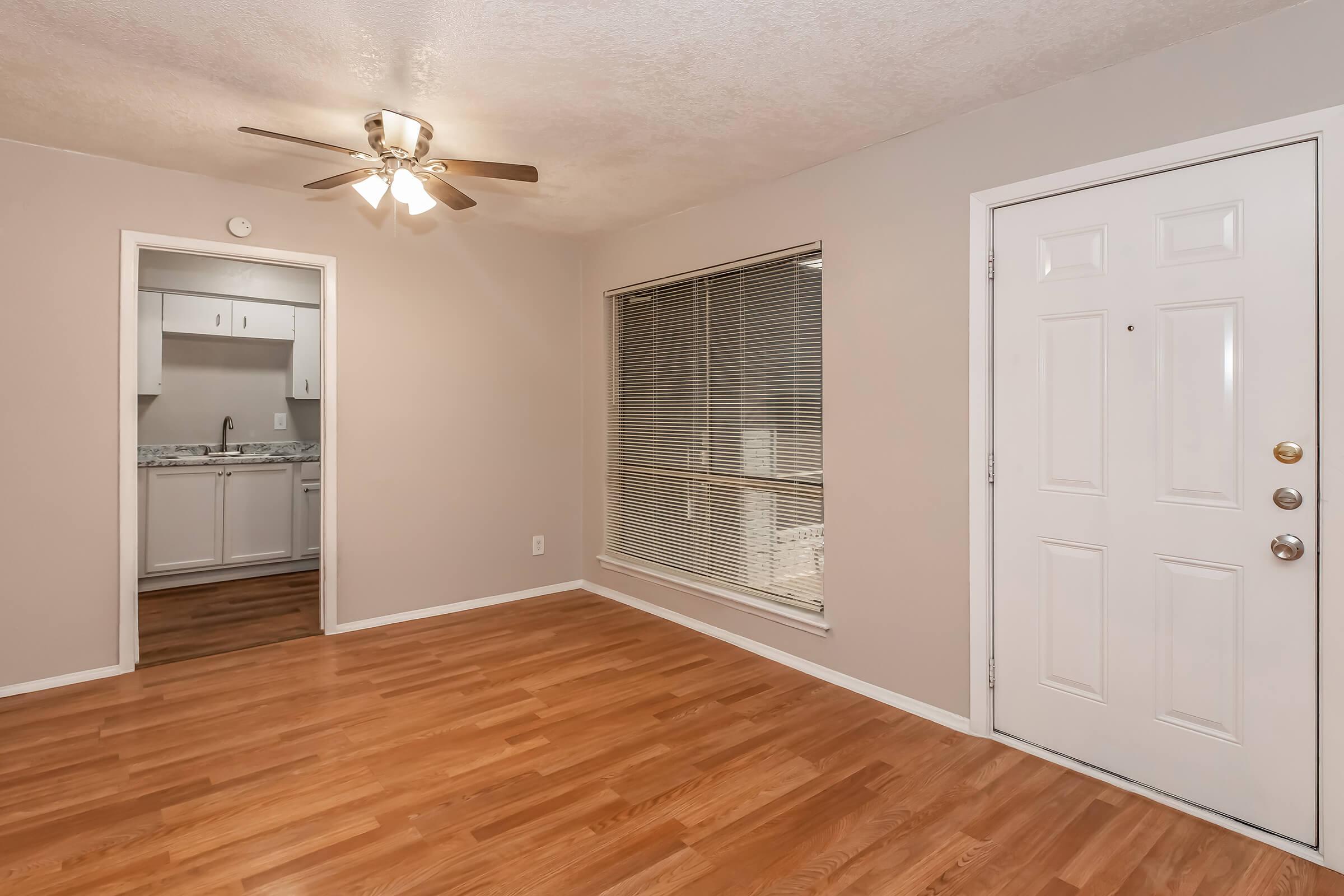 A cozy, well-lit living room featuring light wood flooring, a ceiling fan, and a large window with blinds. A white door leads to the entrance, and a doorway on the left provides access to an adjacent kitchen area with white cabinets and countertops. The walls are painted in a soft beige tone.