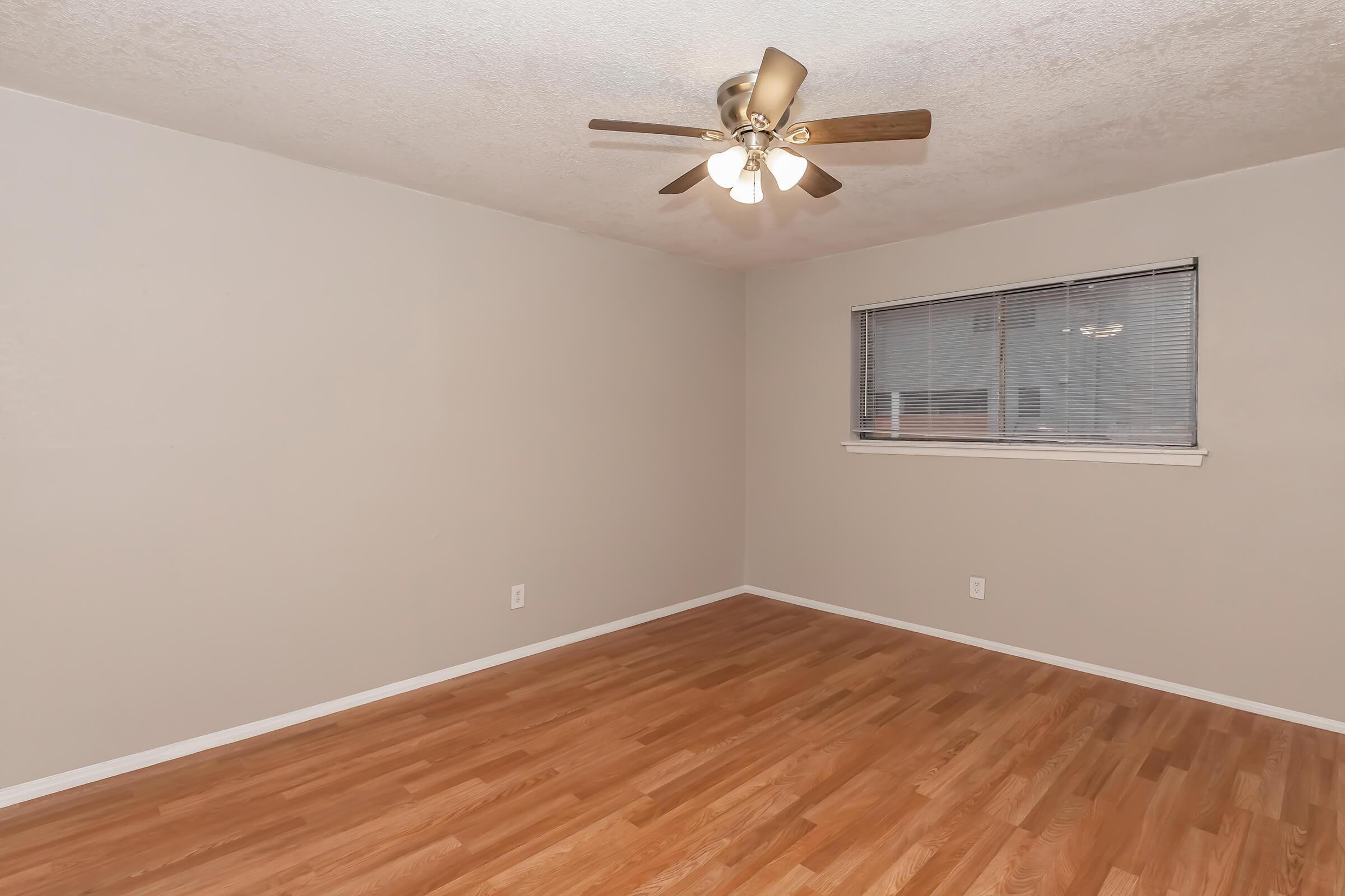 A spacious empty room with light-colored walls and a wooden floor. A ceiling fan with five blades is installed in the center. A window with horizontal blinds allows natural light to enter, illuminating the clean, uncluttered space. The overall atmosphere is bright and inviting.