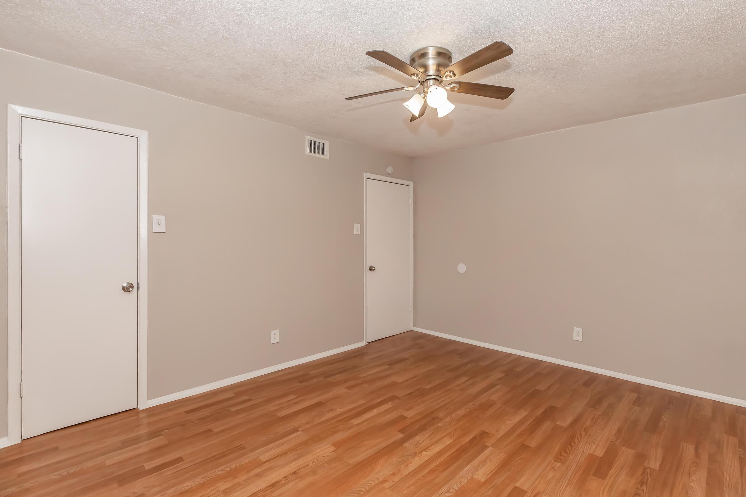 A spacious, empty room with light gray walls and a ceiling fan with four lights. There are two doors: one leading to a closet and the other presumably to another room. The floor is made of light-colored hardwood, enhancing the bright and airy feel of the space.