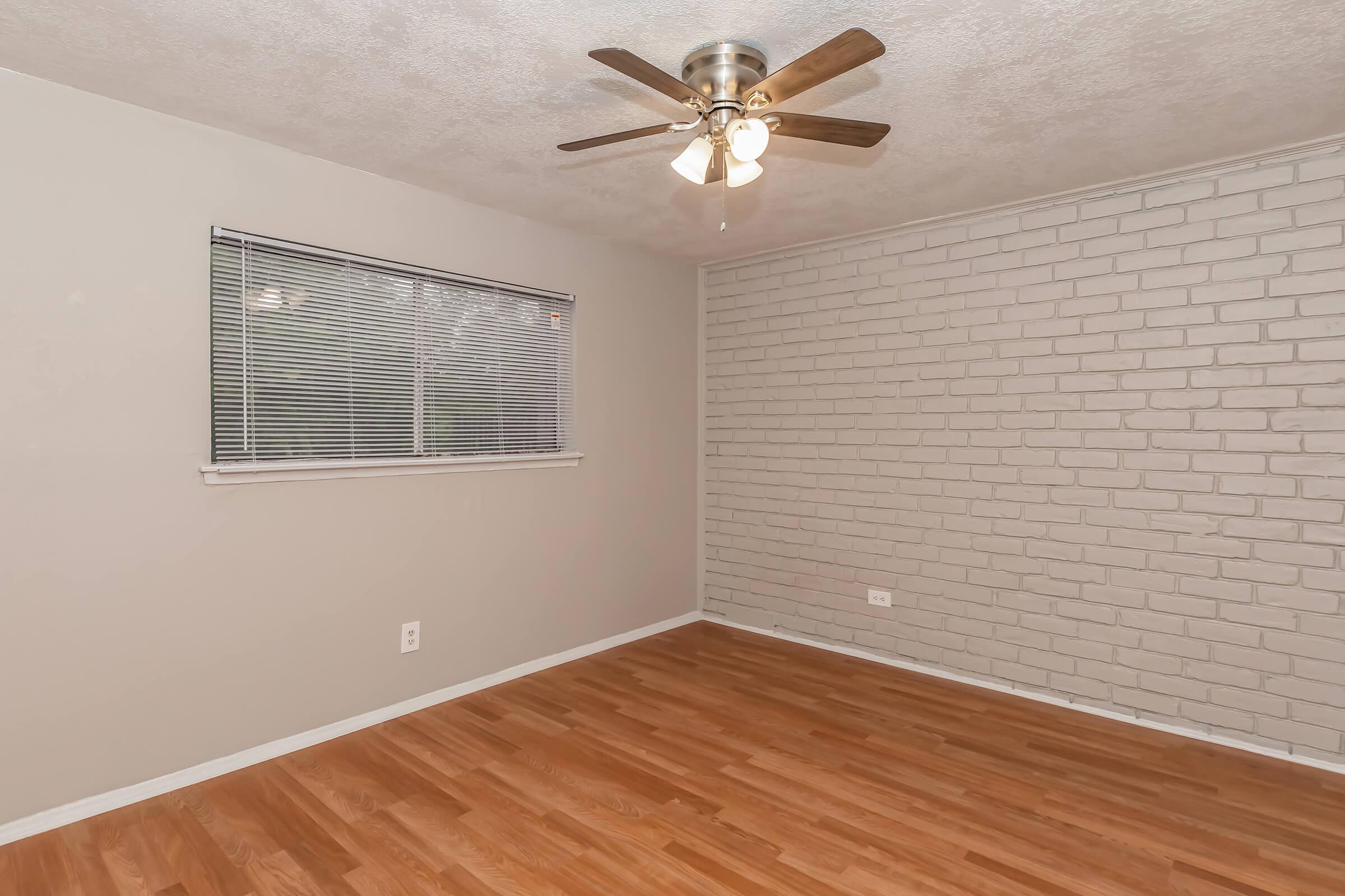 A vacant room featuring a light-colored brick accent wall, a ceiling fan with wooden blades, and a window with blinds. The flooring is made of light-toned wood, and the walls are painted a soft gray, creating a neutral and spacious atmosphere.