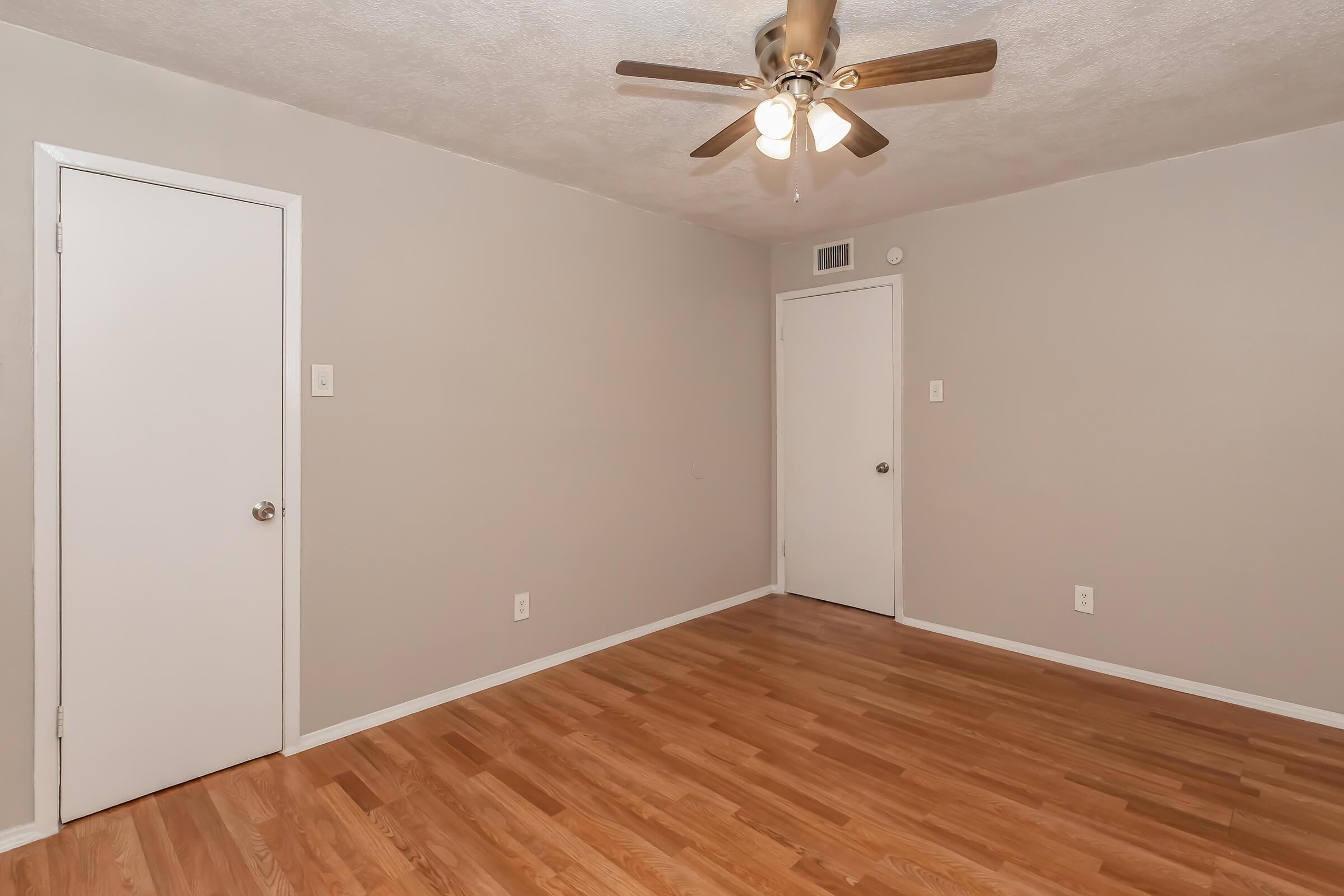 A spacious room featuring light gray walls, a ceiling fan with three bulbs, and hardwood flooring. There are two doors: one leading to another room and another likely to a closet. The design is simple and modern, providing a neutral backdrop for various interior decorating styles.
