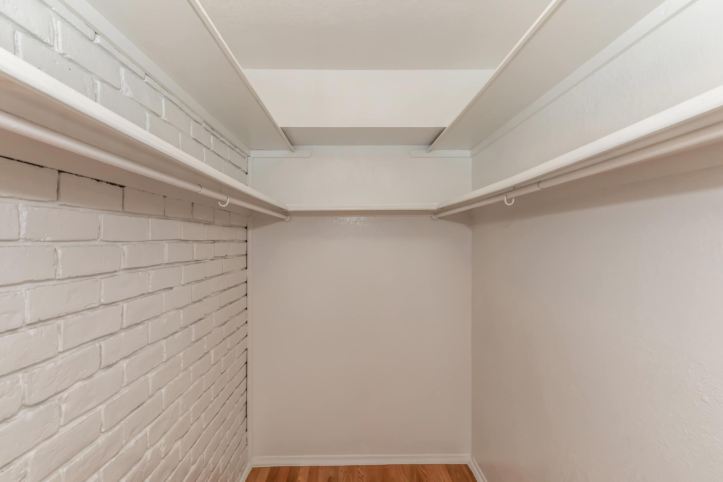 An empty closet with white brick walls and wooden flooring. There are two horizontal metal rods for hanging clothes, and the interior is well-lit with a neutral color scheme. The space appears to be clean and organized, suitable for storage.