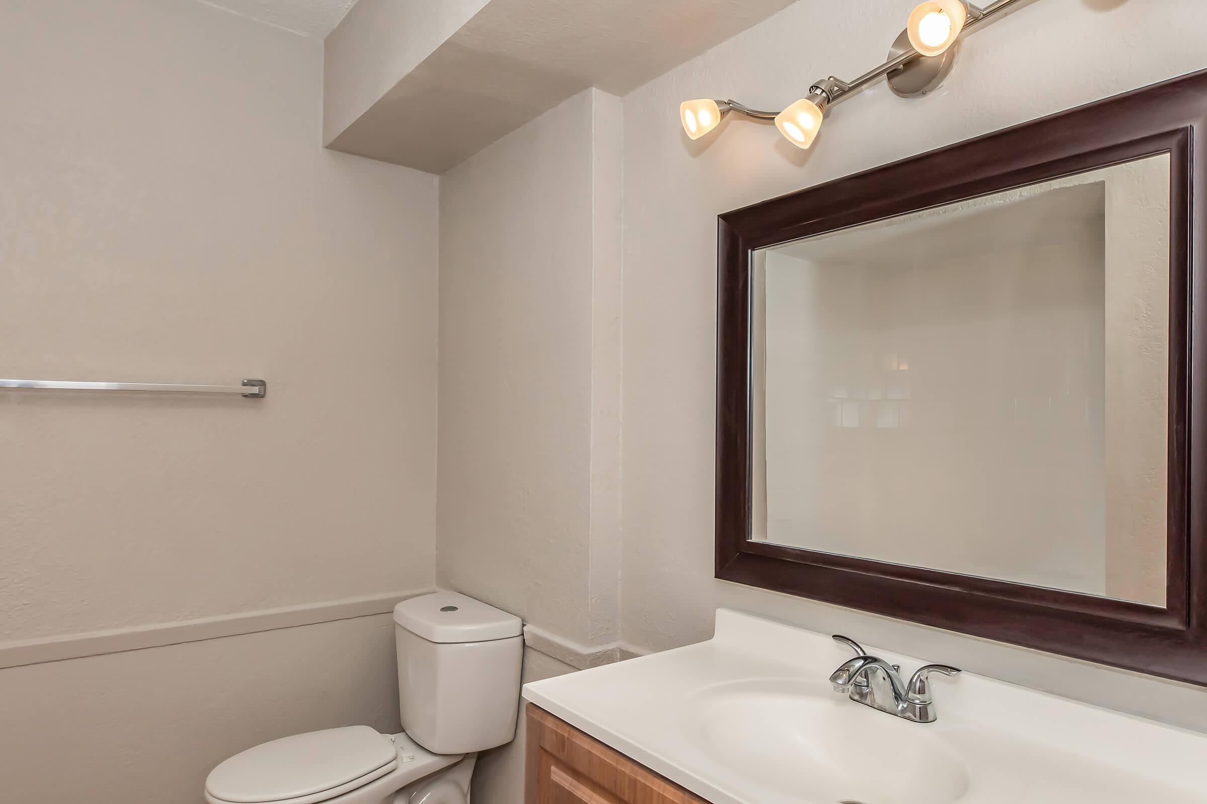 A clean and simple bathroom featuring a toilet, a modern sink with two faucets, a large mirror above the sink, and overhead lighting. The walls are painted in a light color, and there's a towel rack mounted on the wall. Overall, the space has a minimalist and uncluttered design.