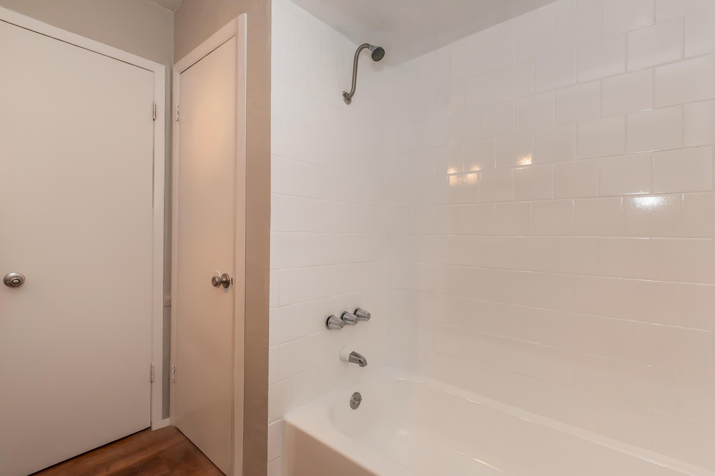 A clean, minimalist bathroom featuring a white tiled wall, a bathtub with metal faucet controls, and a showerhead. A closed white door is visible in the corner, and the floor is a warm wood tone, creating a bright and modern atmosphere.