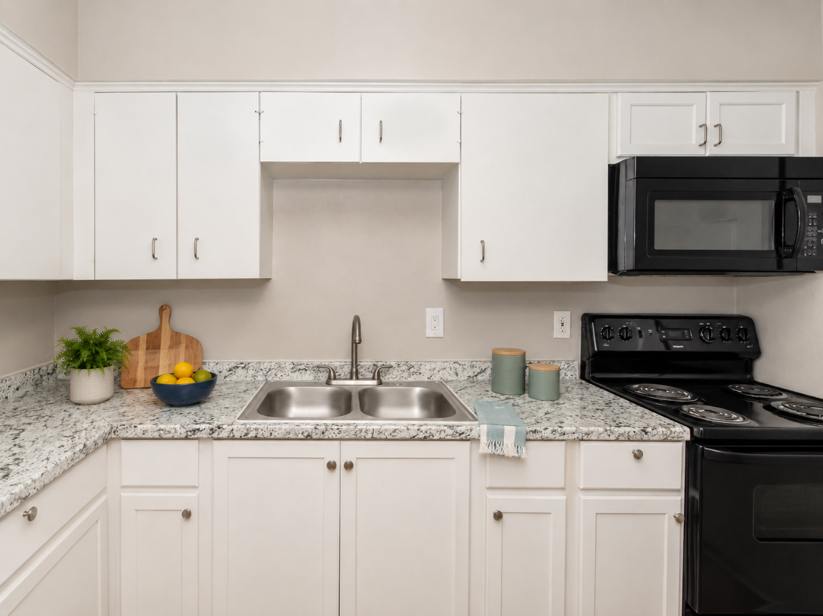 A modern kitchen featuring white cabinetry, a double sink with a chrome faucet, and a granite countertop. The space includes a black microwave and stove, a bowl of lemons and limes, and a potted plant. Soft blue and white kitchen towels are draped by the sink, enhancing the bright and clean aesthetic.