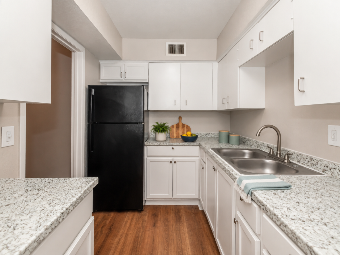 A modern kitchen featuring white cabinets, a black refrigerator, and a gray speckled countertop. There is a double sink and a cutting board with fruit on it. The floor is wooden, and a green plant is placed on the countertop. Natural light brightens the space.
