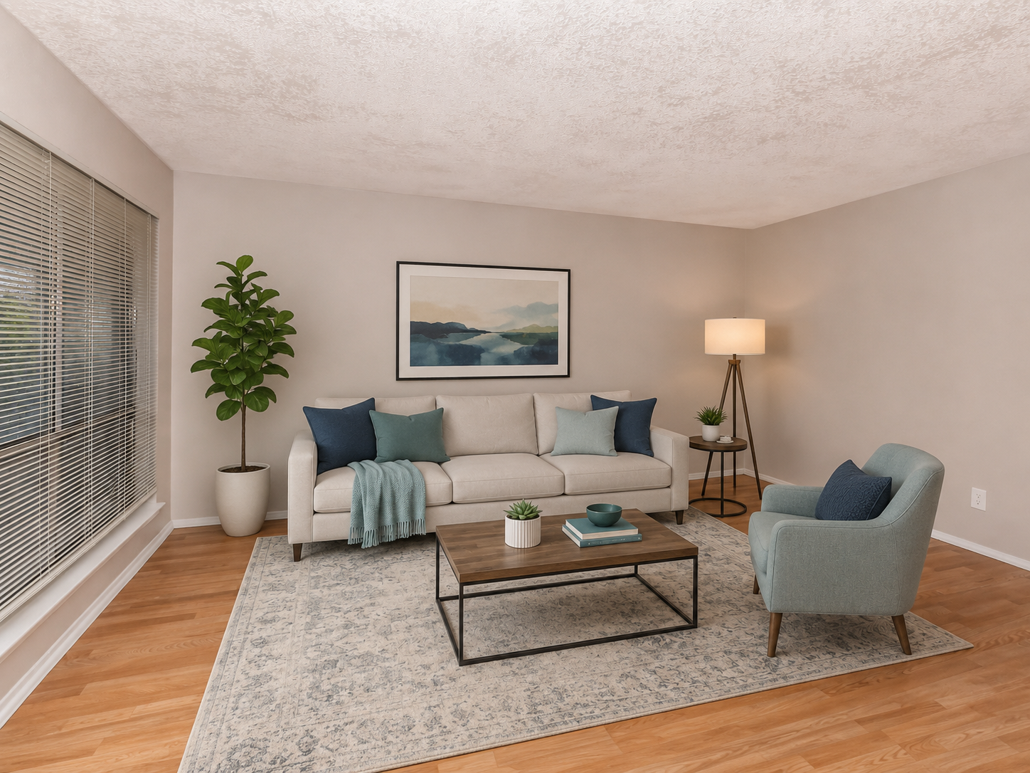 A modern living room featuring a light-colored sofa with blue and teal accent pillows, a small round coffee table, and a cozy armchair. A decorative plant sits in the corner, and a floor lamp provides soft lighting. The backdrop includes a large window with blinds and a framed landscape artwork on the wall.