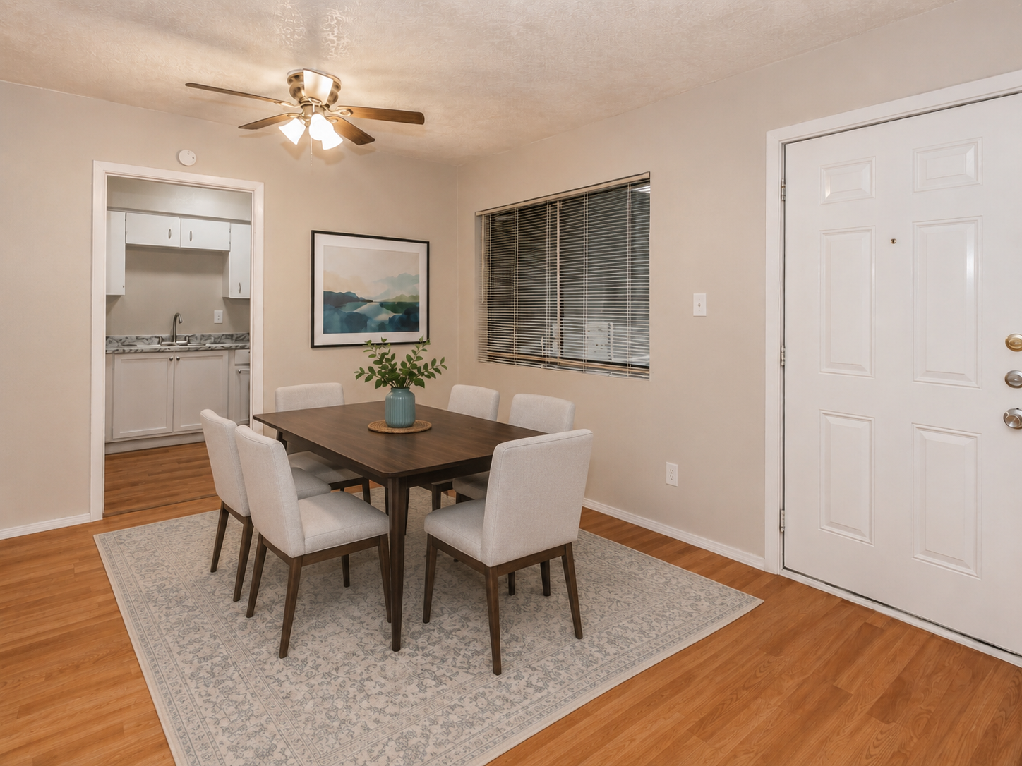 A cozy dining area featuring a wooden table surrounded by six light-colored chairs on a patterned rug. A blue vase with greenery sits on the table. The room has warm lighting from a ceiling fan, and a nearby window hints at an adjacent kitchen with white cabinets. The walls are a soft beige color.