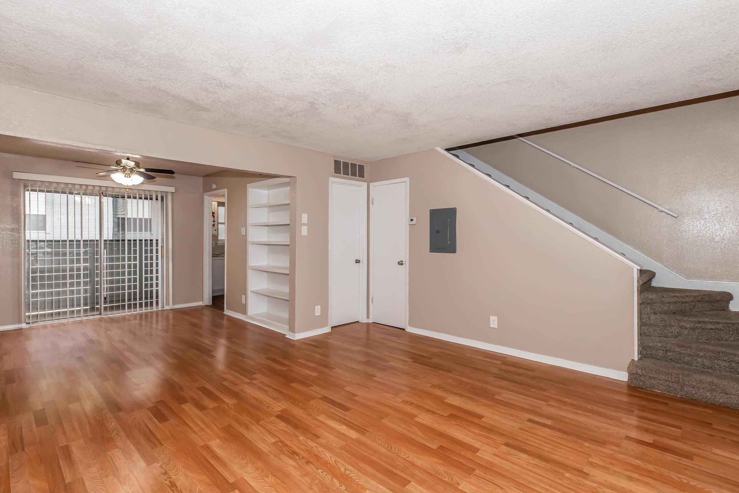 Spacious living room with hardwood floors, ceiling fan, and neutral-colored walls. A staircase is visible on the right, and a sliding glass door leads to a balcony with vertical blinds. A small nook with built-in shelves is on the left, and a closed door indicates access to other areas of the home.