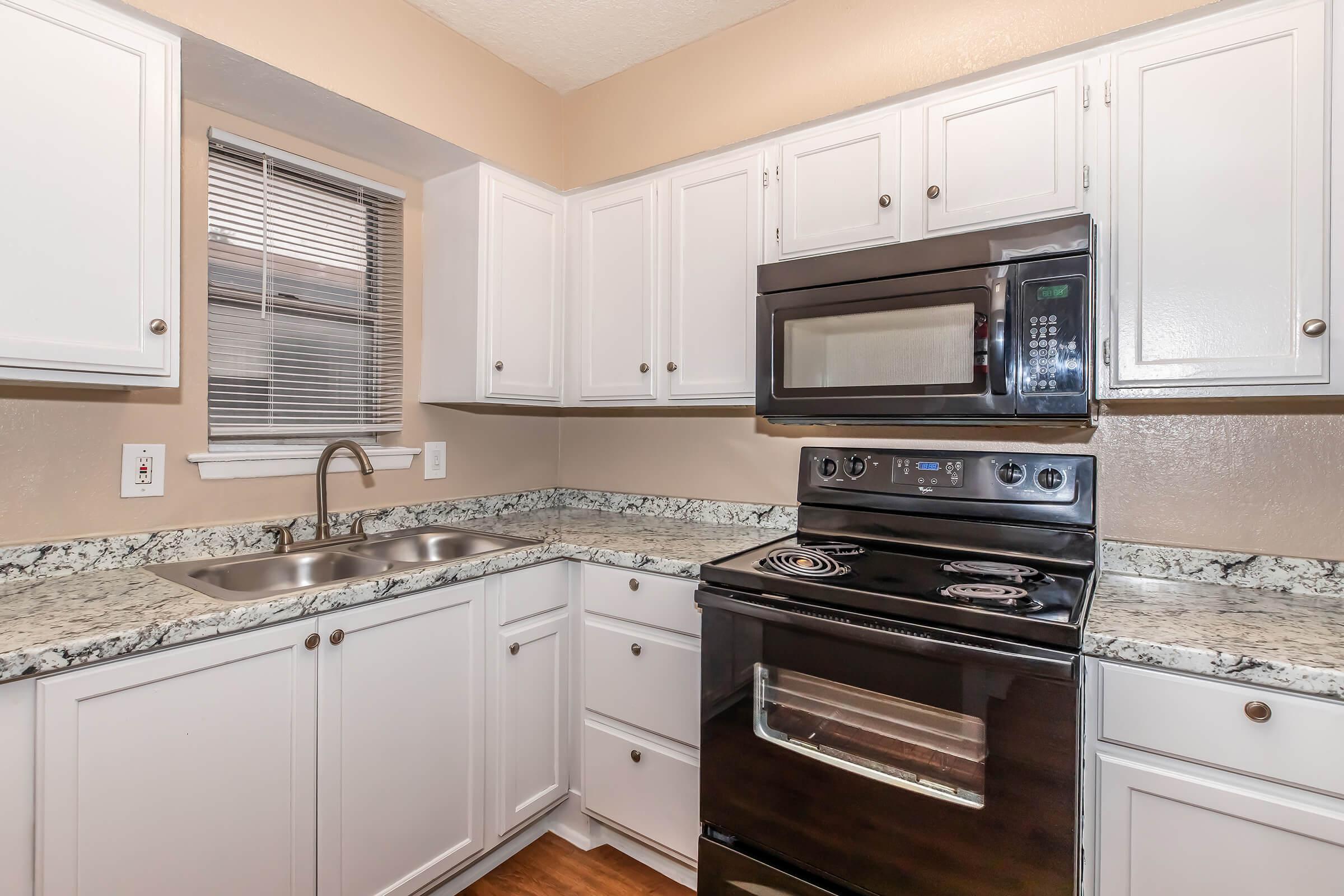A modern kitchen featuring white cabinetry, a double sink with a brushed nickel faucet, a black microwave above a black electric stove, and a granite countertop. Natural light enters through a window with light-colored blinds, and the floor has a warm wooden finish, creating a cozy and inviting atmosphere.