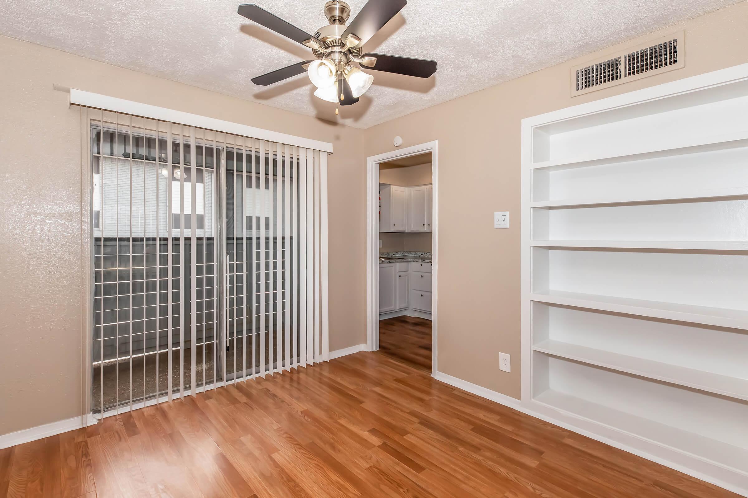 A cozy interior living area featuring hardwood flooring, a ceiling fan with light fixtures, and a sliding glass door leading to a balcony. The walls are painted a neutral color, and there is a built-in white bookshelf against one wall. A doorway opens into a kitchen area.