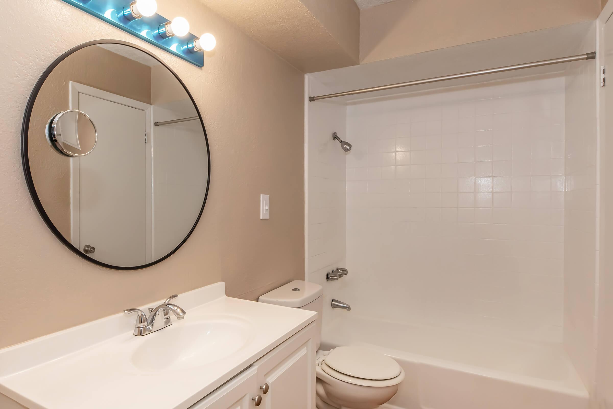 A clean, modern bathroom featuring a white tiled bathtub with a showerhead, a round mirror above a white vanity with a sink, and two light fixtures above the mirror. The walls are painted in a warm neutral tone, adding to the inviting atmosphere. A compact toilet is located next to the bathtub.