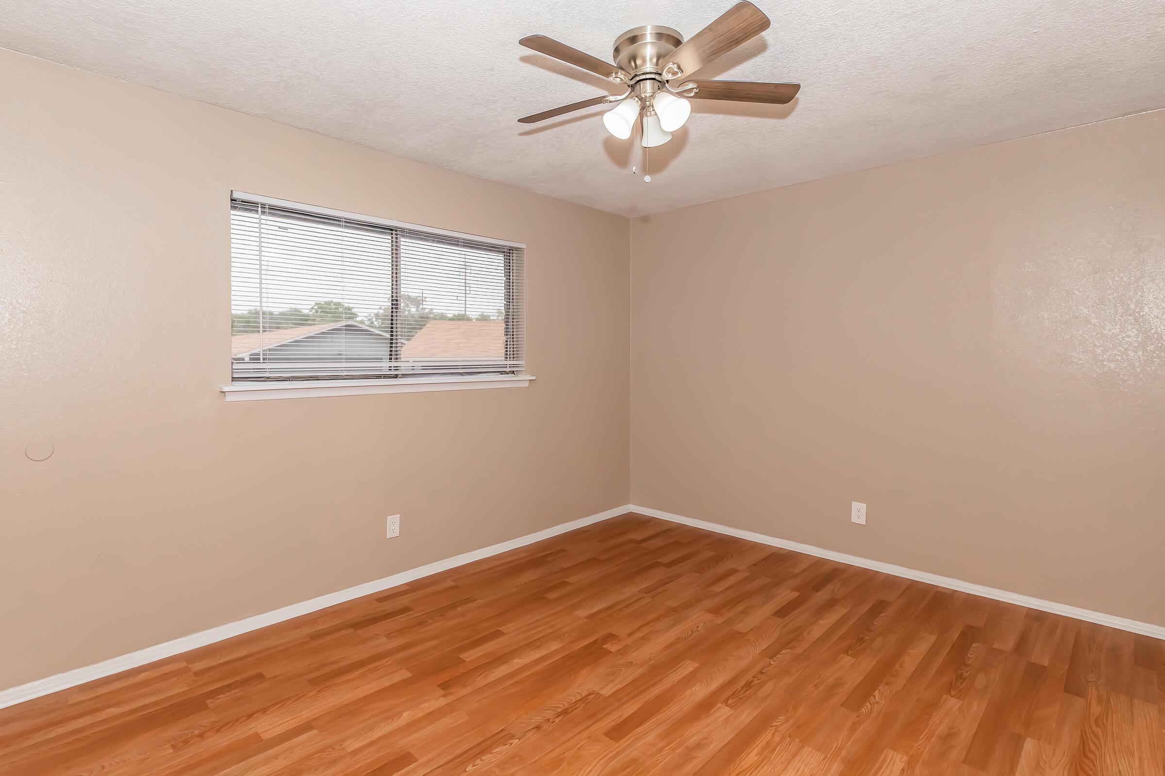 A light beige room with a wooden floor, featuring a ceiling fan with three lights. There is a window on the left side with blinds, allowing natural light to enter. The walls are smooth and painted in a matching beige color, creating a bright and inviting atmosphere.