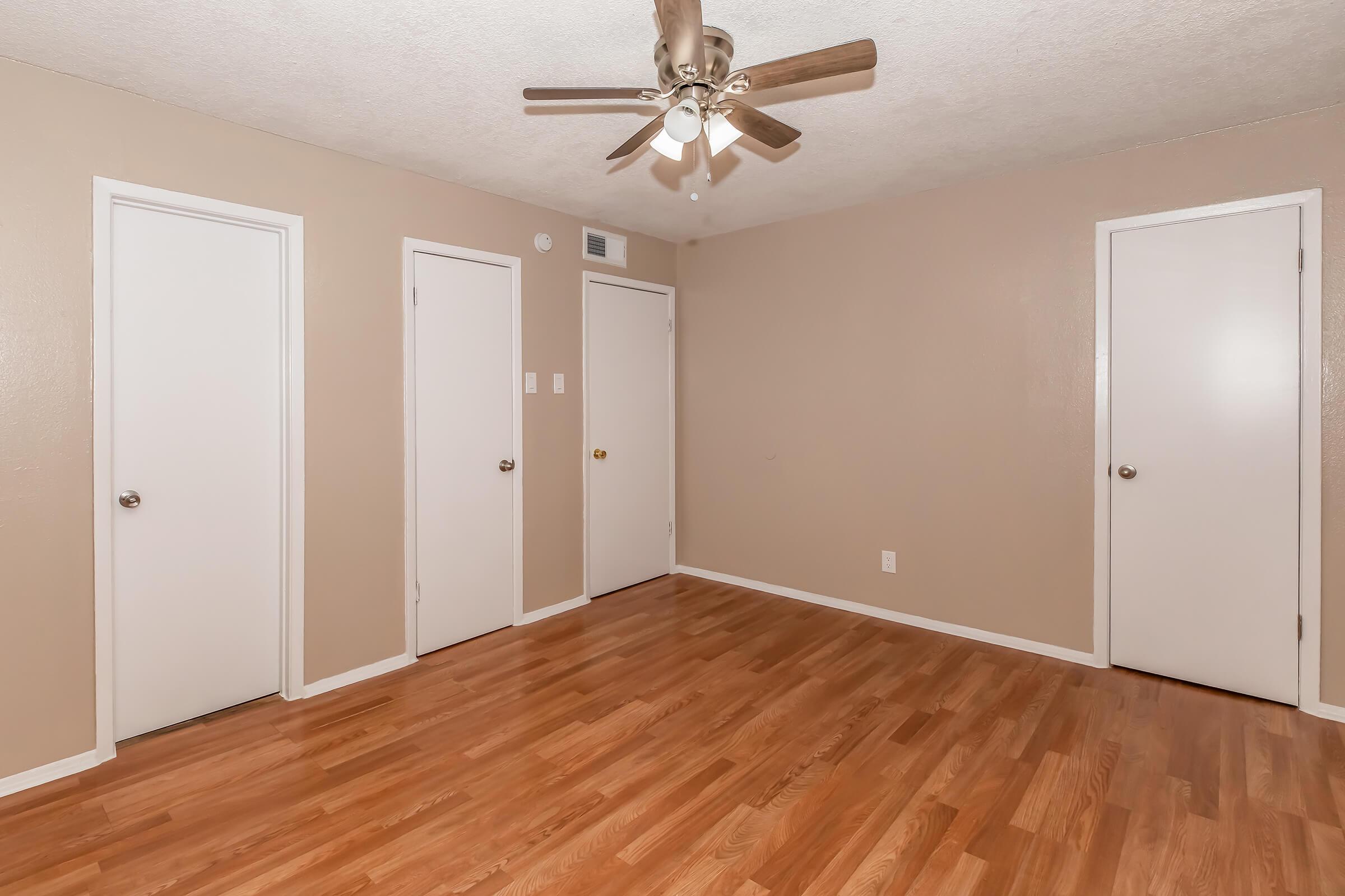 A simple, empty room with light brown walls and hardwood flooring. The ceiling features a ceiling fan with light fixtures, and there are three white doors on the walls. The room appears spacious and well-lit, showcasing a clean and minimalistic design.