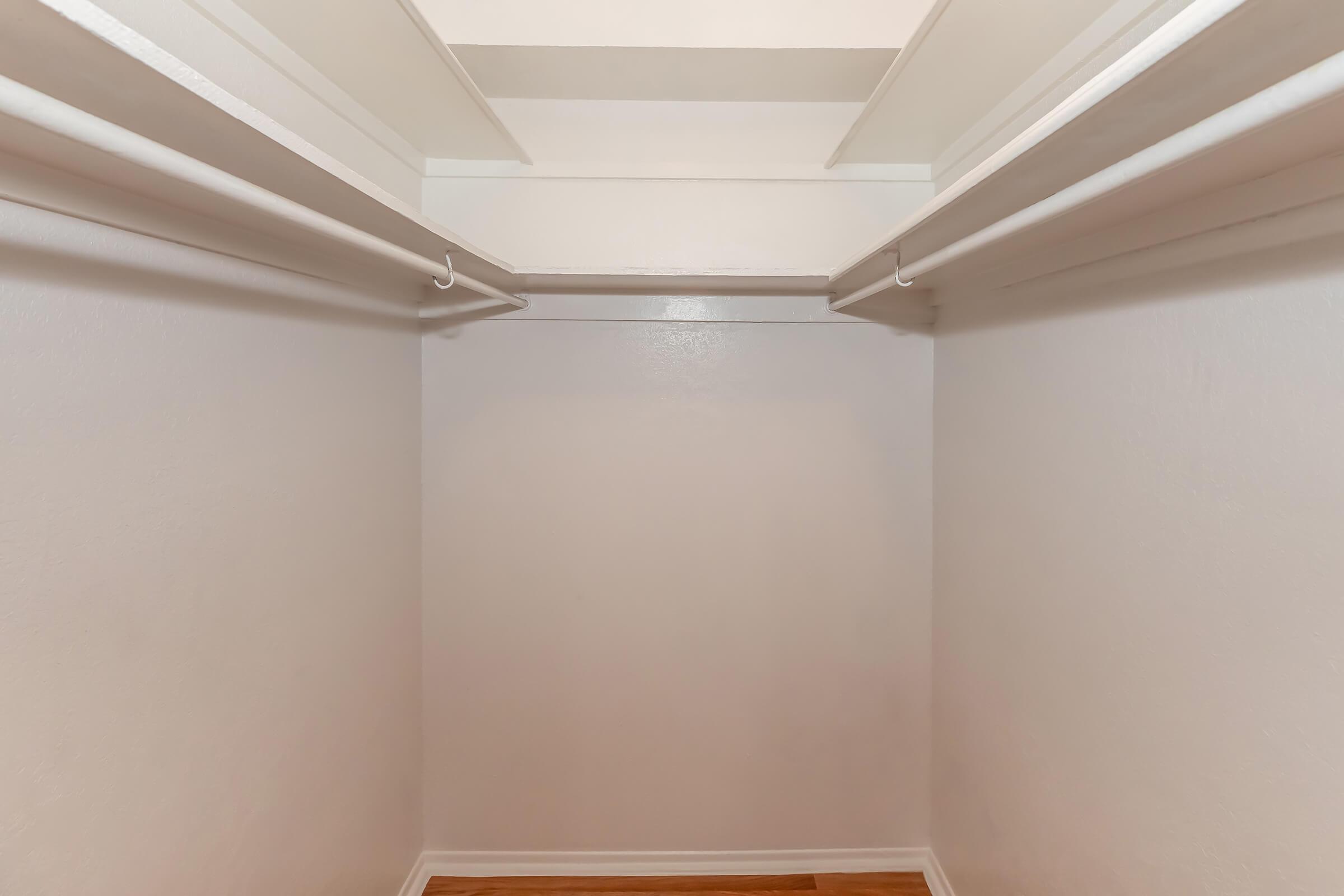 Empty closet interior with two hanging rods on white walls and wooden flooring. The space is well-lit and minimalistic, lacking any items or shelves, creating a clean and open appearance.