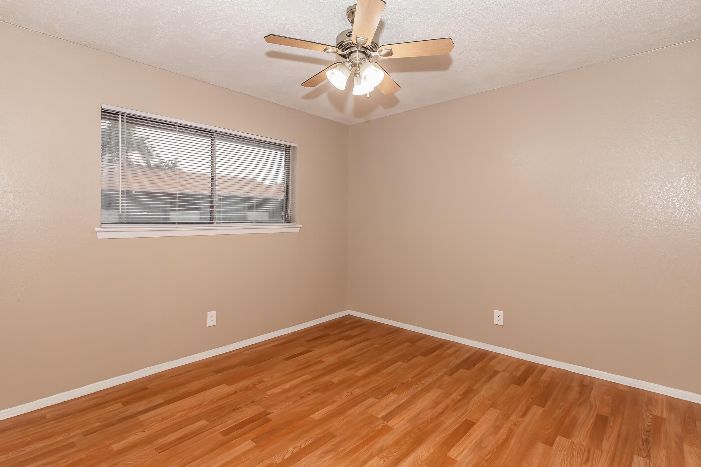 A spacious room with wooden flooring, a ceiling fan with four lights, and a window with blinds. The walls are painted a neutral beige color, and there are no furniture or decorations visible, creating an empty and clean atmosphere.