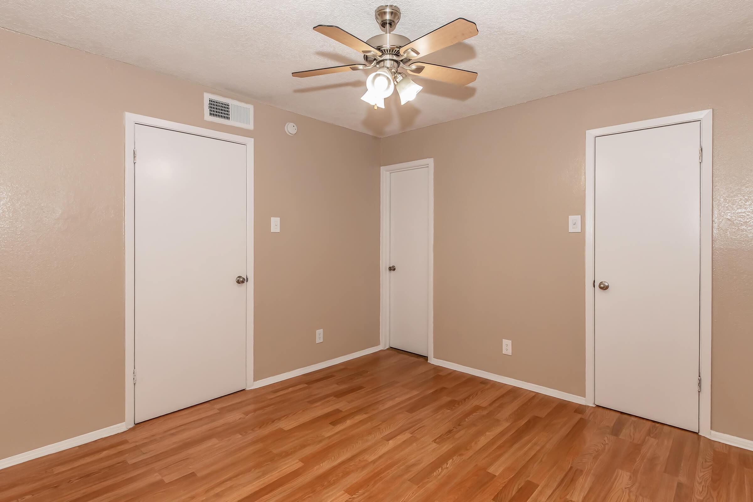 A corner of a room with light brown walls and a ceiling fan. There are two closed doors on the walls, one on the left and one on the right. The floor is made of light-colored hardwood, creating a warm and inviting atmosphere. The room appears spacious and well-lit.