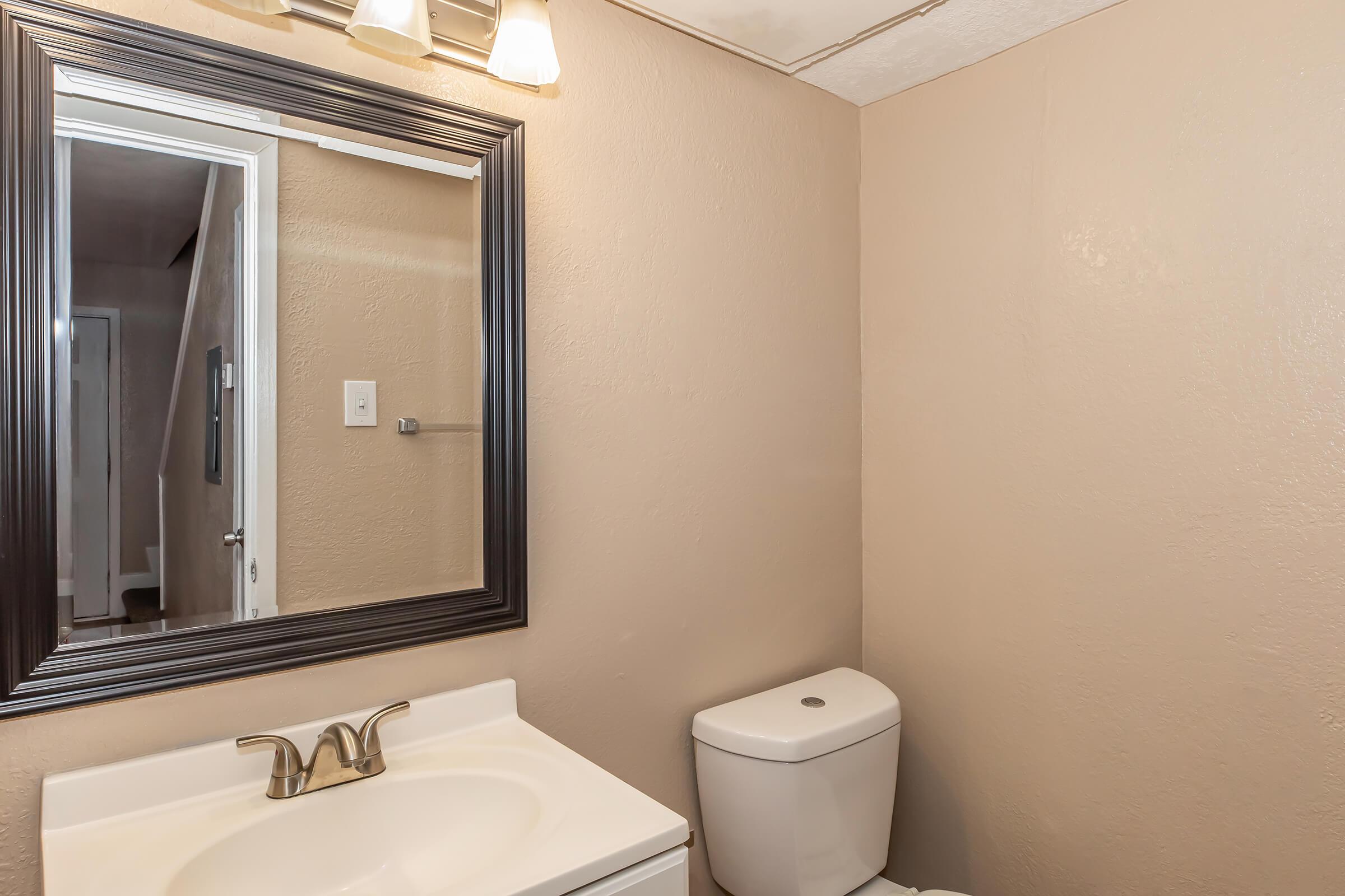 A well-lit bathroom featuring a white countertop sink with a single faucet, a modern toilet, and a large framed mirror above the sink. The walls are painted in a light beige color, and a partially visible door leads to another room, creating a clean and tidy appearance.