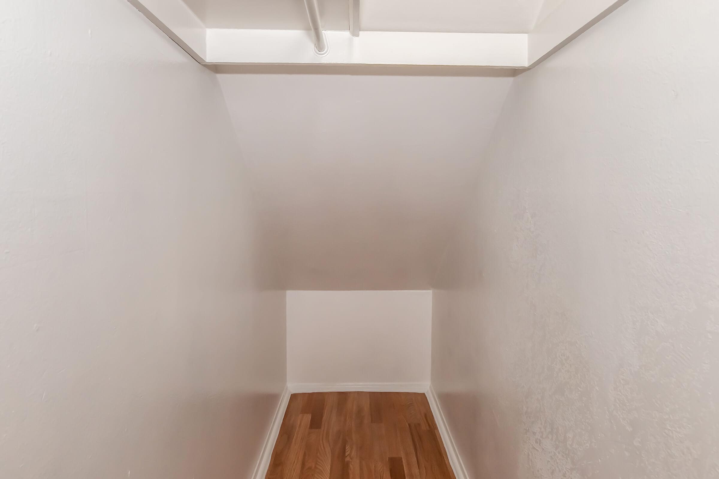 A small, empty room with sloped white walls and a wooden floor. The space has minimal lighting and is sparsely furnished, creating a clean and open feel. A ceiling fixture is visible at the top, adding a touch of illumination to the otherwise plain area.