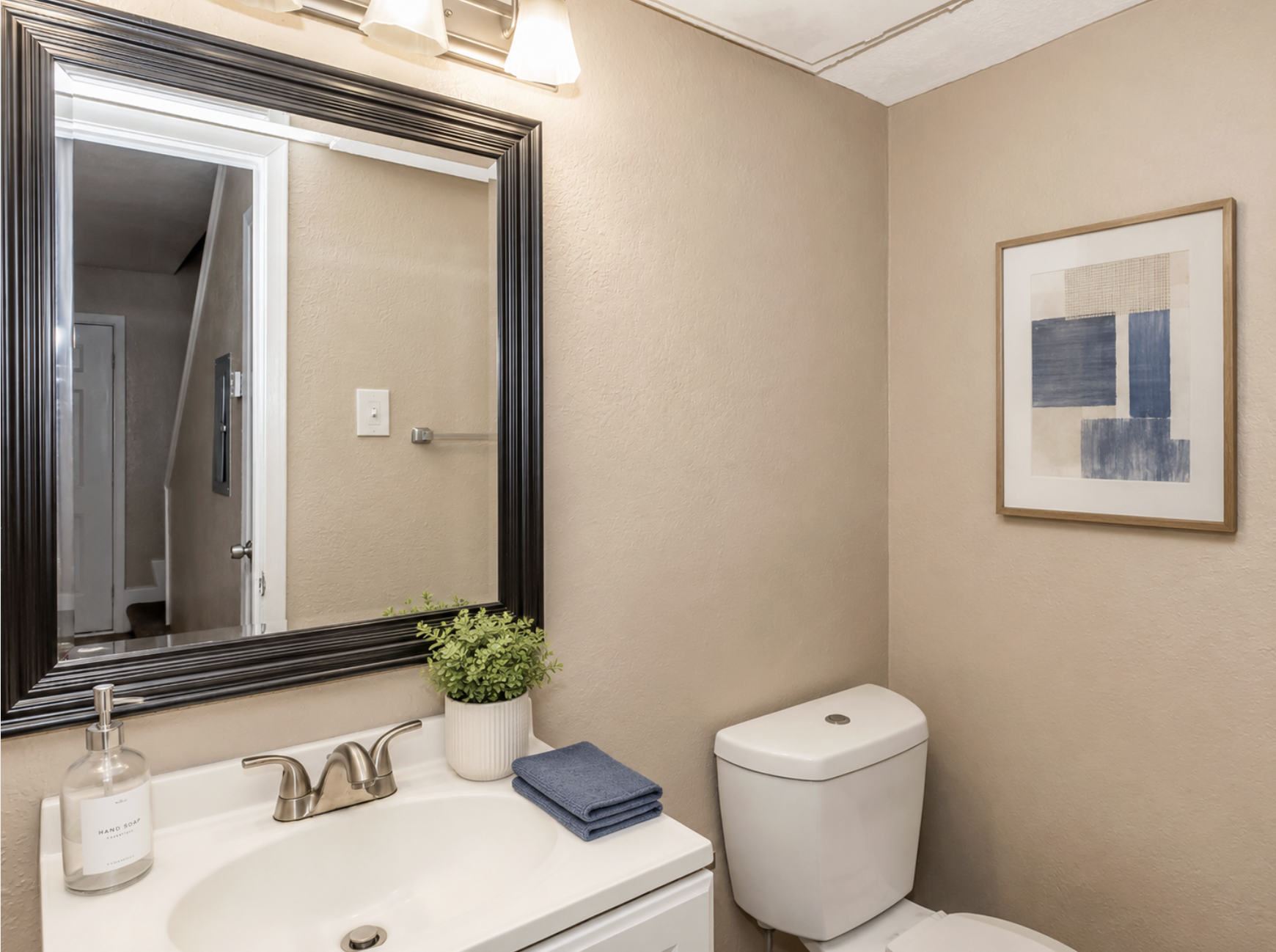 A modern bathroom featuring a white vanity with a sink, a large mirror framed in black, and a stylish wall art piece. The toilet is positioned next to it, and a small plant adds a touch of greenery. Soft blue towels are neatly placed on the countertop, complementing the warm beige walls.