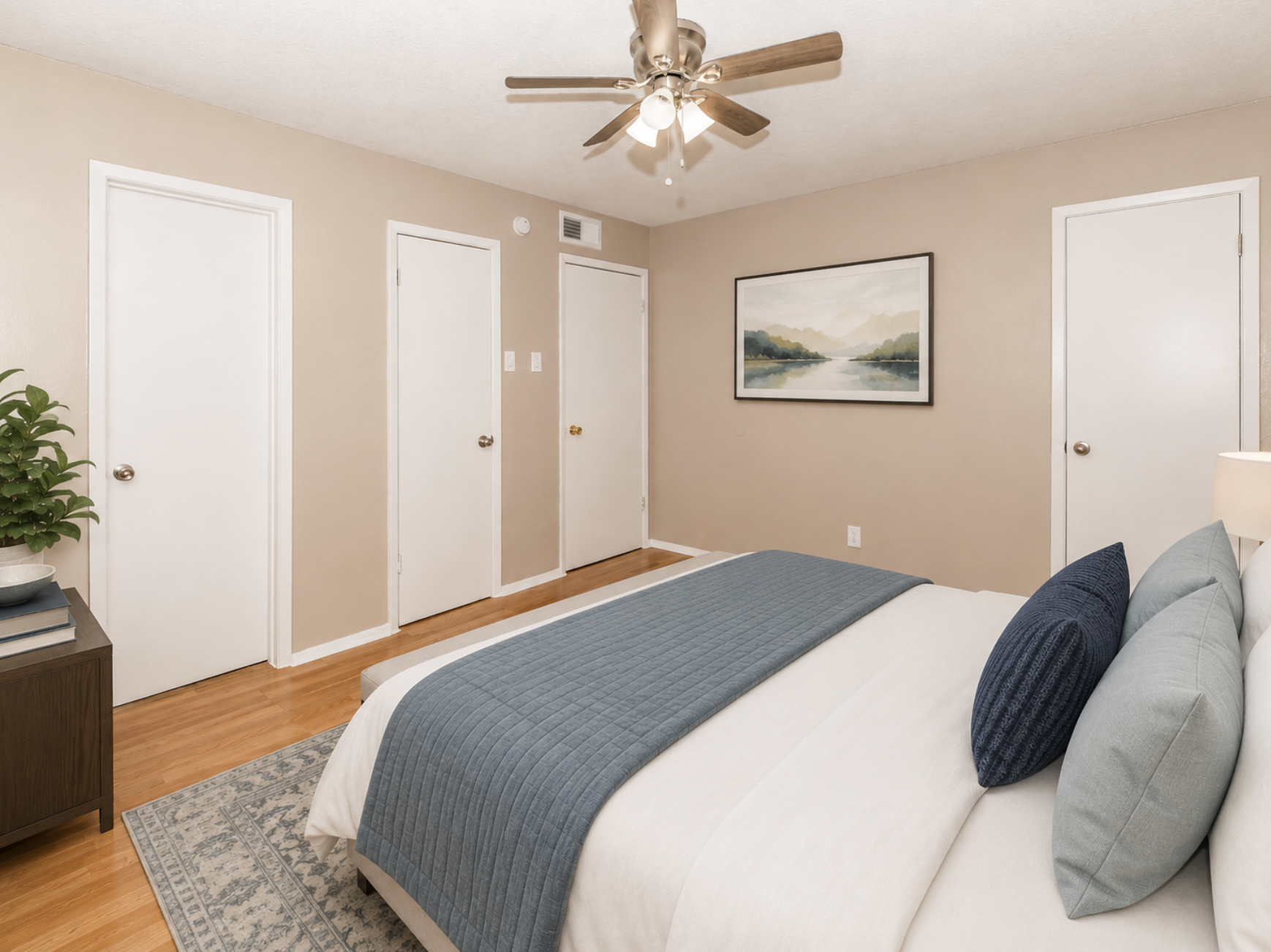 A cozy bedroom featuring a queen-sized bed with a blue throw blanket and two decorative pillows. The room has beige walls and hardwood flooring, with a ceiling fan and two closed white doors. A piece of artwork depicting a landscape hangs above a nightstand with a potted plant. Natural light fills the space.