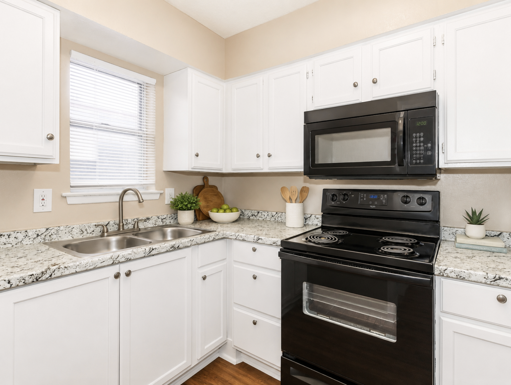 A stylish kitchen with white cabinets, granite countertops, and a black stove and microwave. There is a stainless steel sink below a window, wooden utensils in a holder, green apples in a bowl, and small plants on the counter, creating a bright and inviting space.