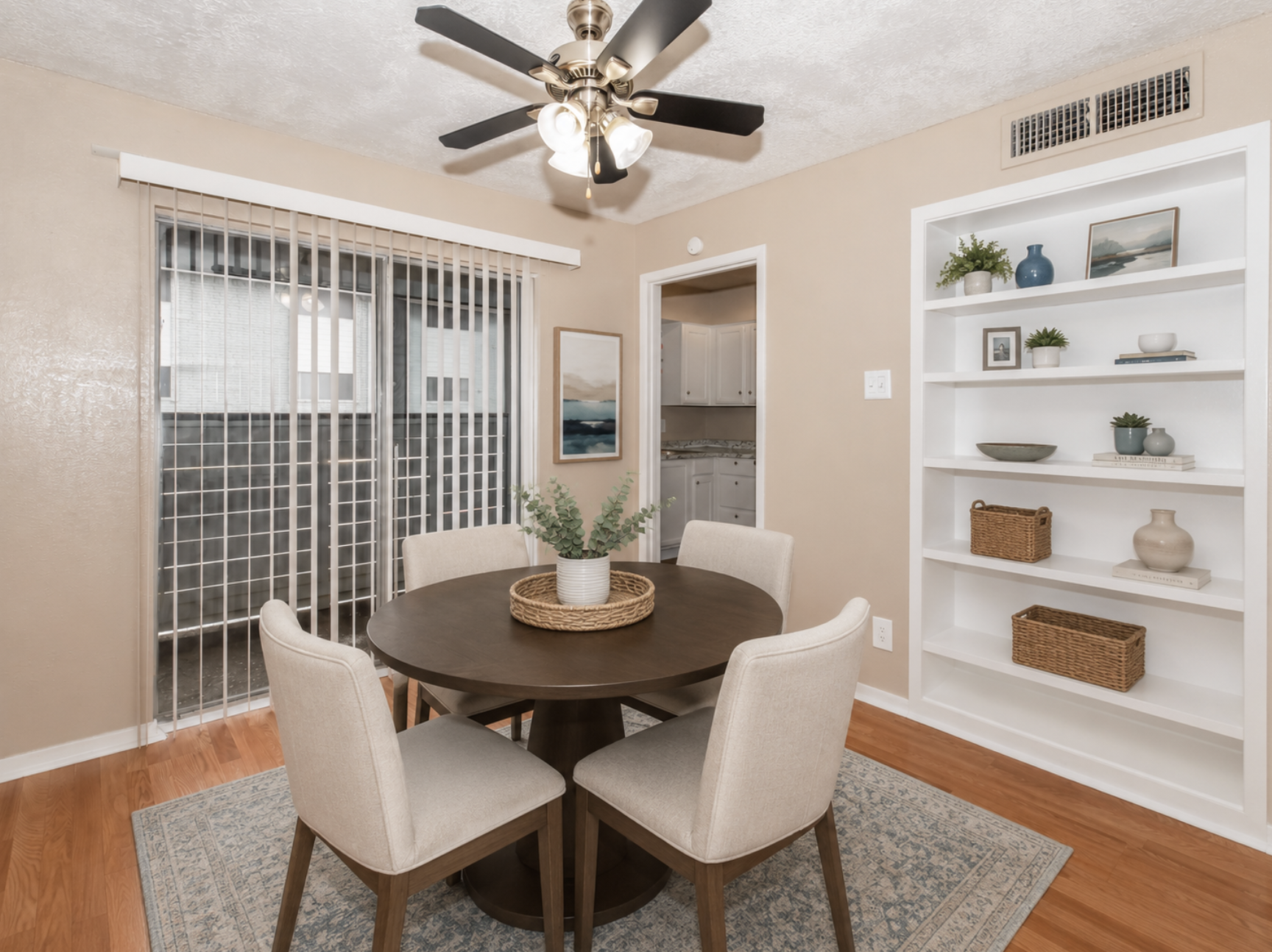 A cozy dining area featuring a round wooden table surrounded by four light-colored chairs. A ceiling fan with light fixtures hangs above. The walls are painted a warm beige, and a sliding door with vertical blinds provides natural light. A white shelf displays decorative items and plants.