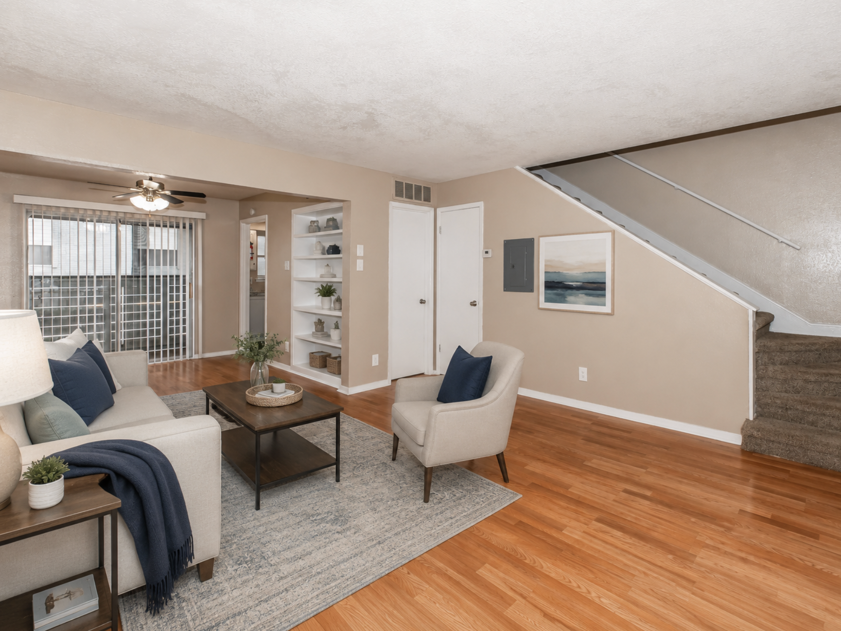 A cozy living room featuring a neutral sofa with a blue throw, an armchair, and a wooden coffee table. Light walls and hardwood flooring create a warm atmosphere. A staircase is visible in the background, along with a shelf displaying decor. Large windows allow natural light to fill the space.