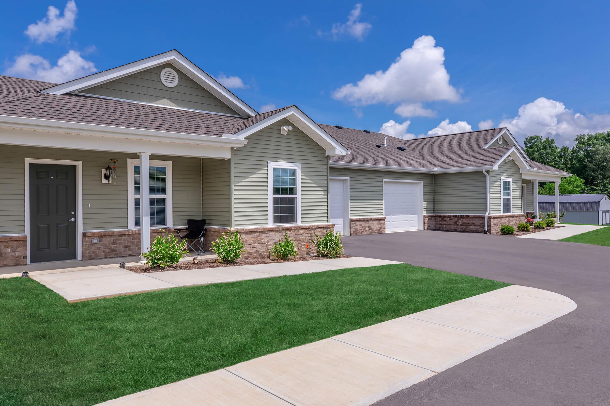 A well-maintained residential property featuring a light green house with a dark front door, a paved driveway, and a garage. The front yard has neatly trimmed grass and landscaped edges. The background includes a bright blue sky with fluffy white clouds.