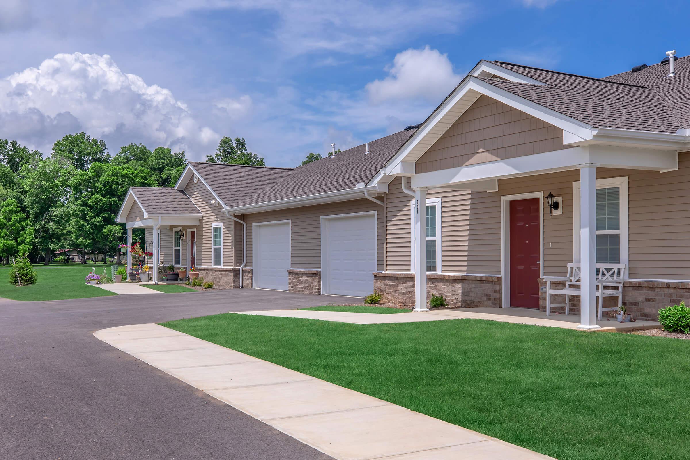A row of modern single-story homes with light brown siding and white trim. Each home features a front porch and garage. The pathway leading to the doors is lined with green grass, and the sky above is partly cloudy, creating a peaceful neighborhood atmosphere.