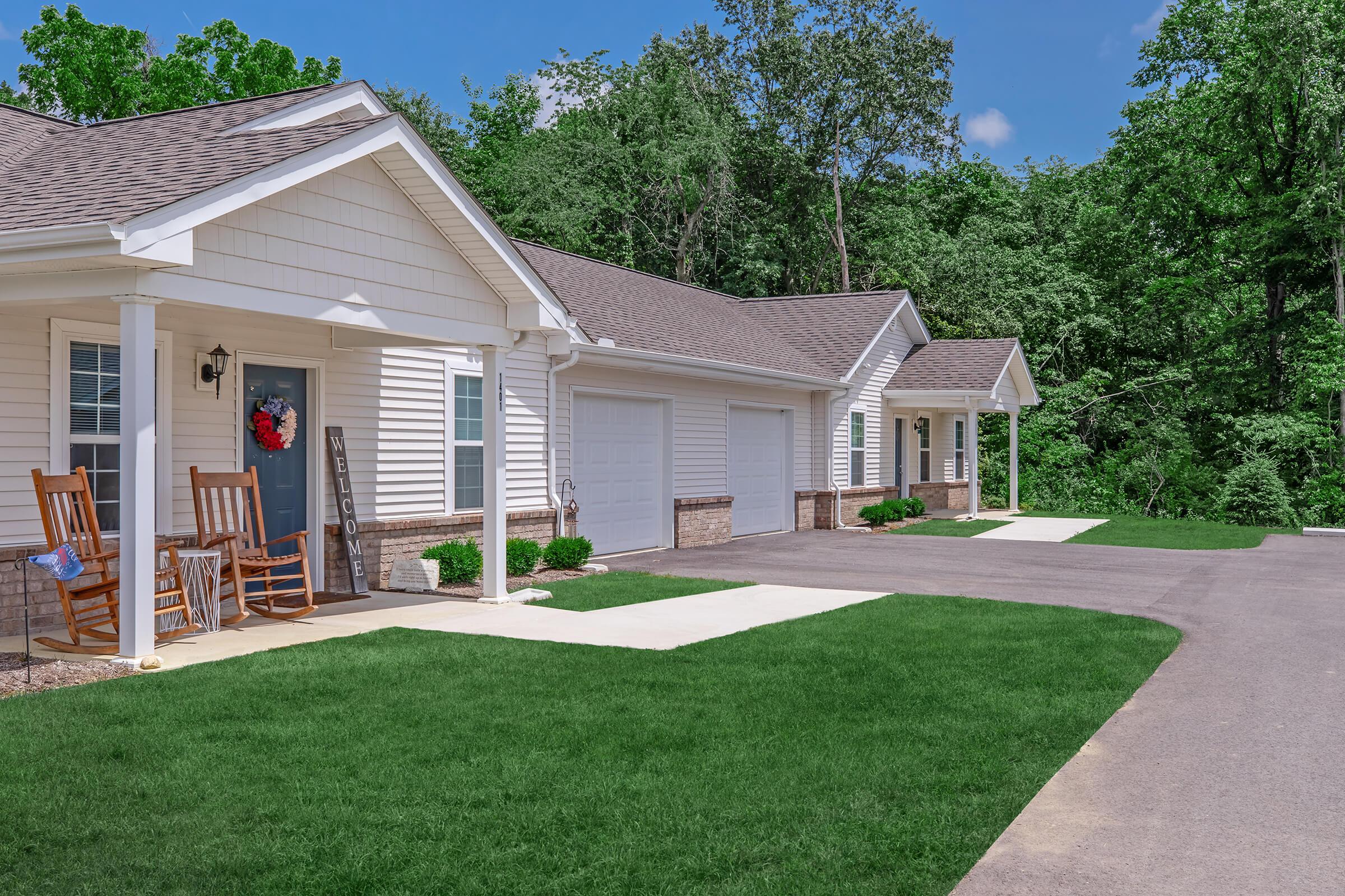A row of charming single-story houses with white siding and beige accents, featuring covered porches and rocking chairs. The well-maintained front lawns are green and manicured, with a paved driveway leading to the garages. Lush trees and blue skies enhance the serene atmosphere of the neighborhood.