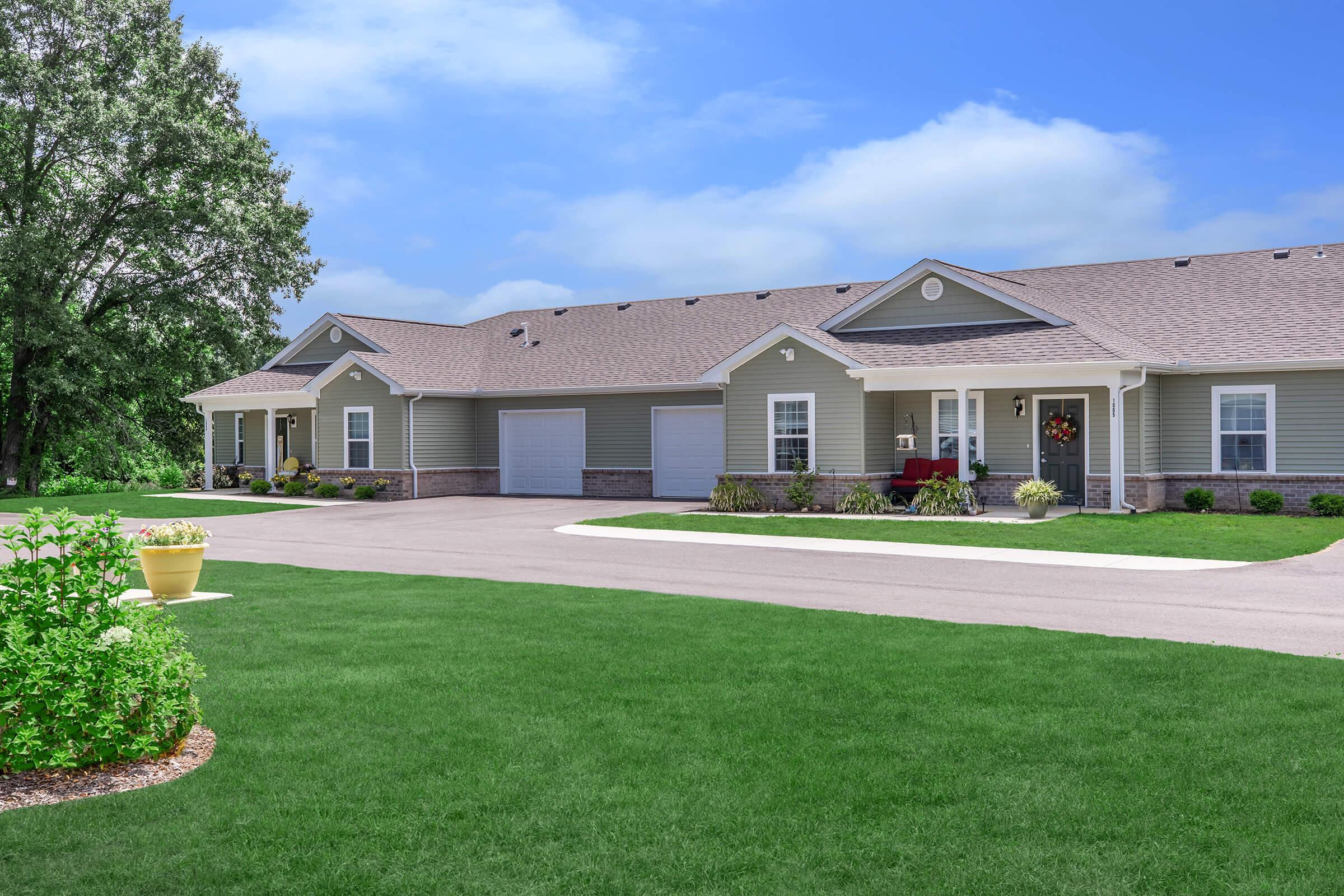 A well-maintained single-story residential building with a gray exterior and a pitched roof. The entrance features a welcoming porch with a decorative wreath. Lush green lawns and flower beds enhance the surroundings, and a paved driveway leads to a garage. The sky is clear with a few clouds.
