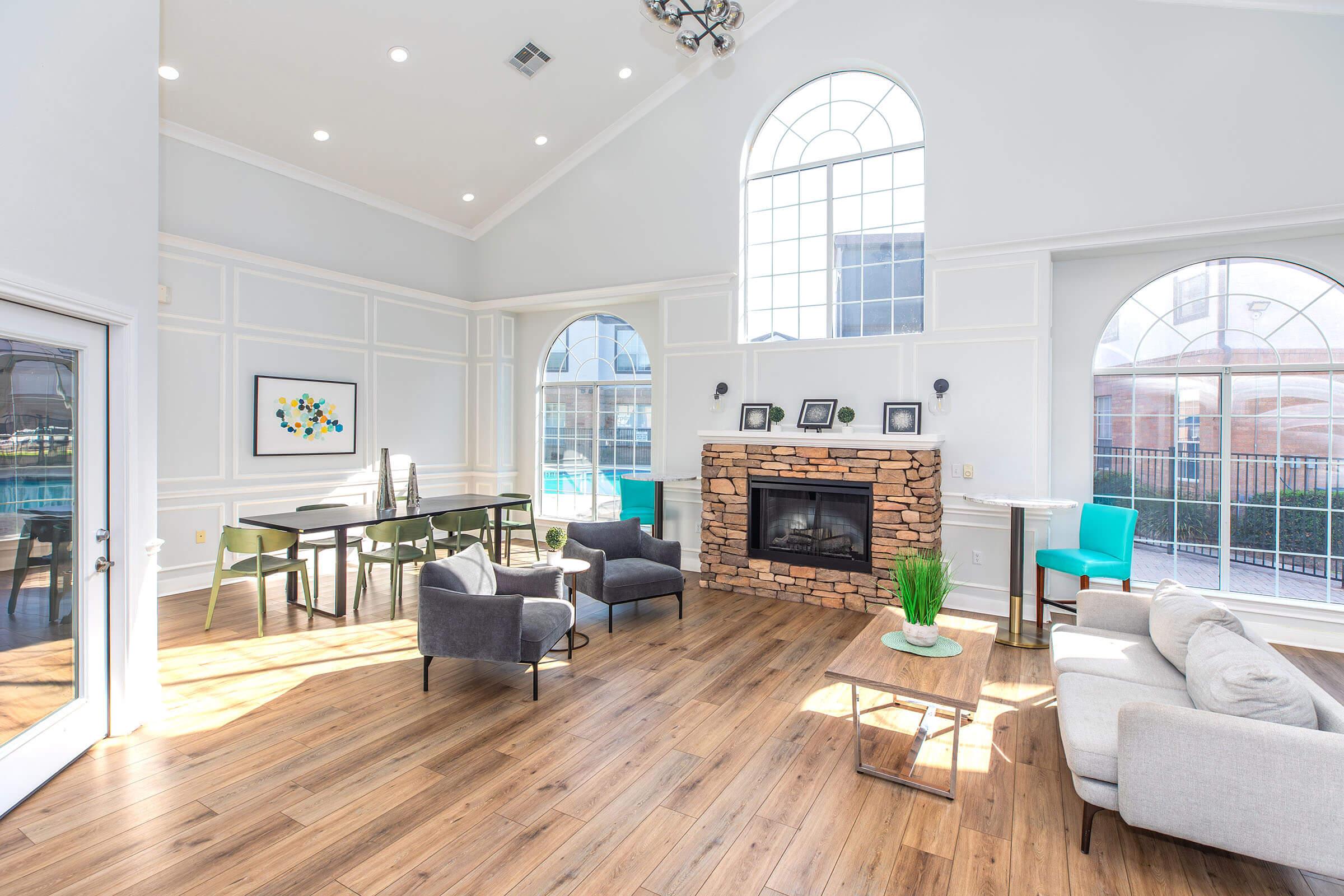 Brightly lit modern living space featuring large arched windows, a stone fireplace, and a mix of seating options, including gray chairs and a light sofa. A wooden table and chairs are situated in the background. The flooring is wood, complementing the contemporary decor with a green plant as a focal point.