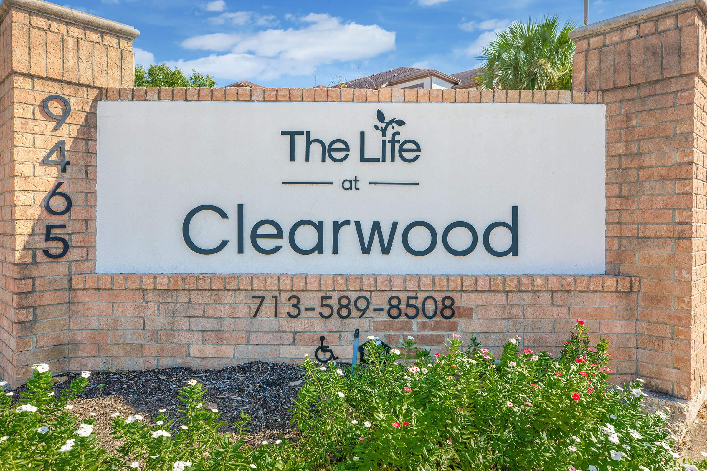 Sign for "The Life at Clearwood," featuring the address 9465 and phone number 713-589-8508. The sign is prominently displayed with brick surrounds and landscaped plants in front. The background includes a blue sky with some clouds.