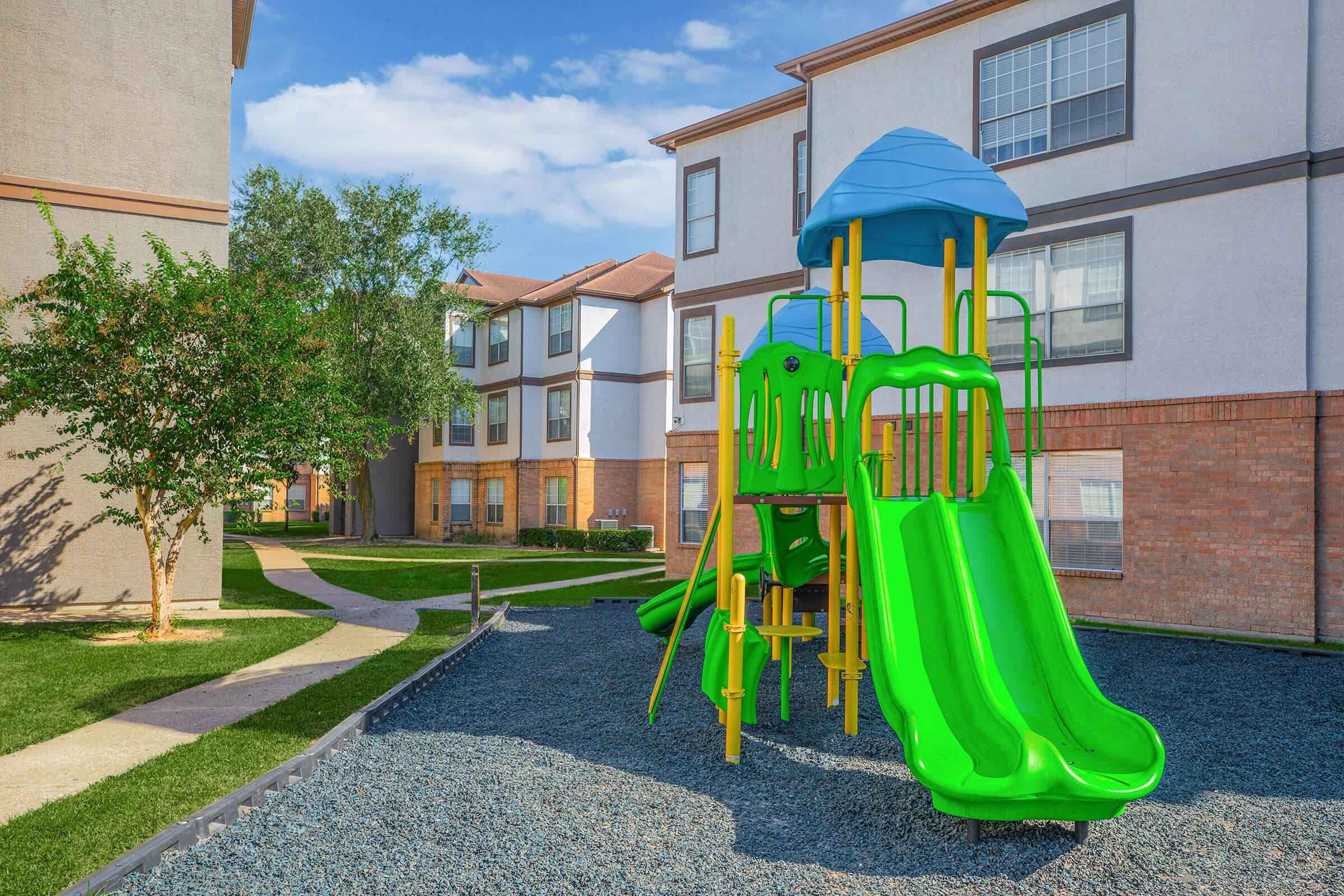 Colorful playground equipment featuring a green slide and climbing structure, situated on a gravel surface in front of residential buildings. The area is landscaped with grass and trees, under a blue sky with scattered clouds.