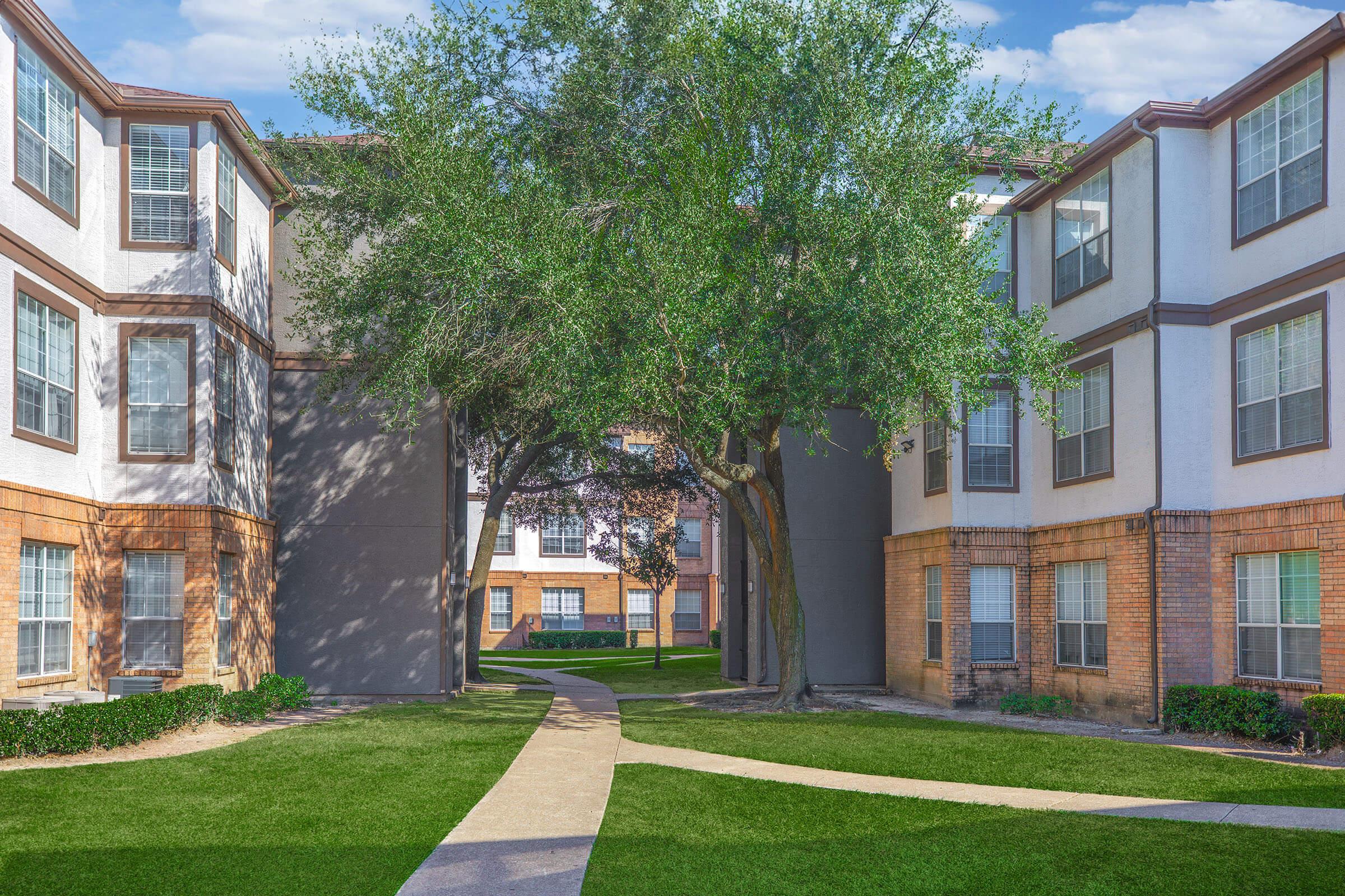 A pathway curves between two apartment buildings with large windows, surrounded by green grass and trees. The scene is bright and sunny, creating a welcoming atmosphere in a residential area.
