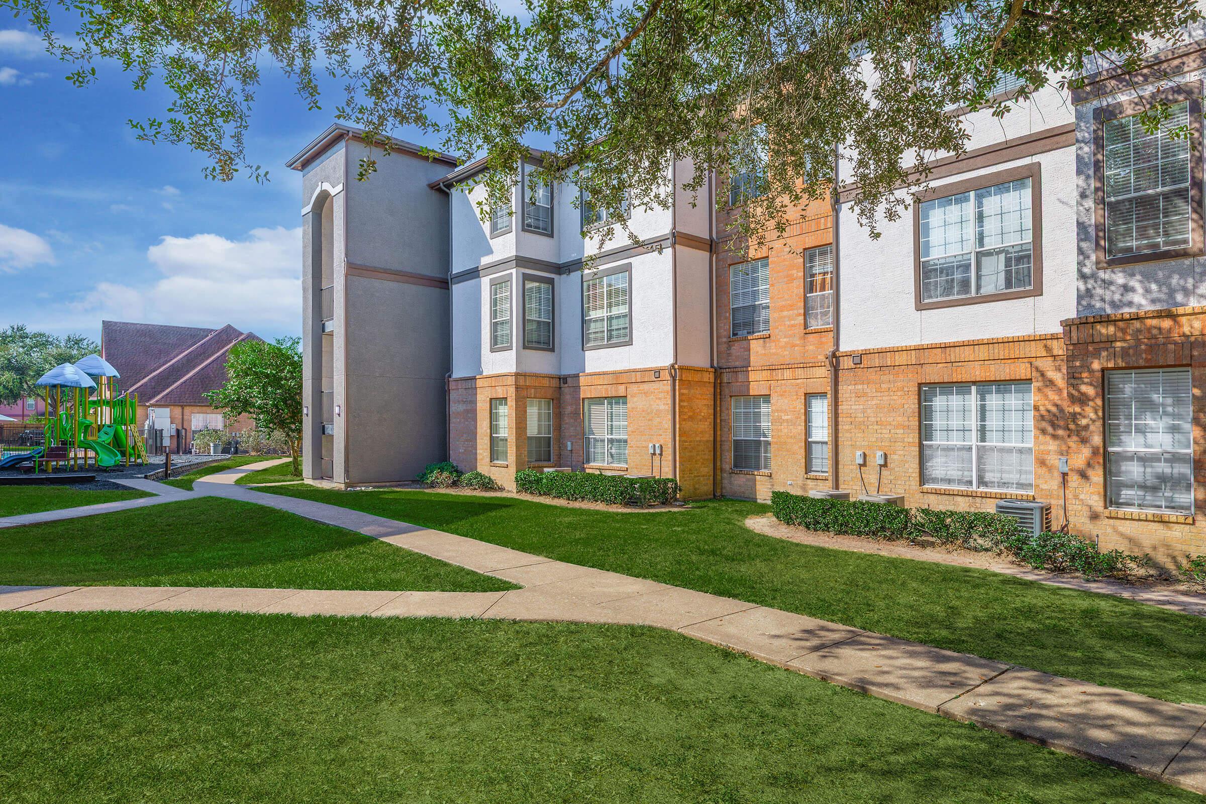 A well-maintained apartment complex featuring a mix of brick and stucco façades. The landscaped area includes neatly trimmed grass, walking paths, and a playground in the background, surrounded by trees and blue skies. The overall scene conveys a welcoming community atmosphere.